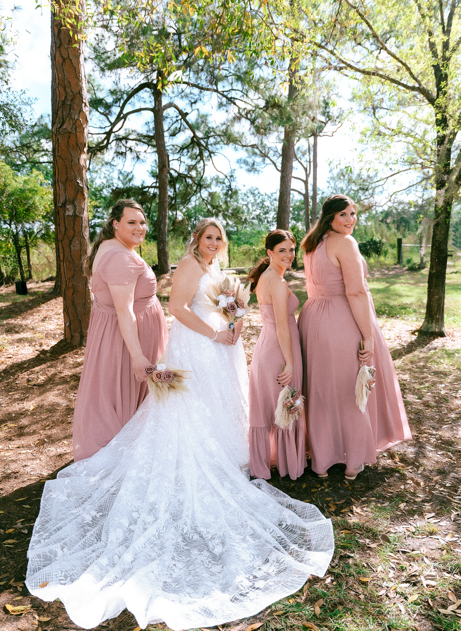 Bride in white lace gown poses with three bridesmaids in dusty rose dresses, holding bouquets outdoors among pine trees.