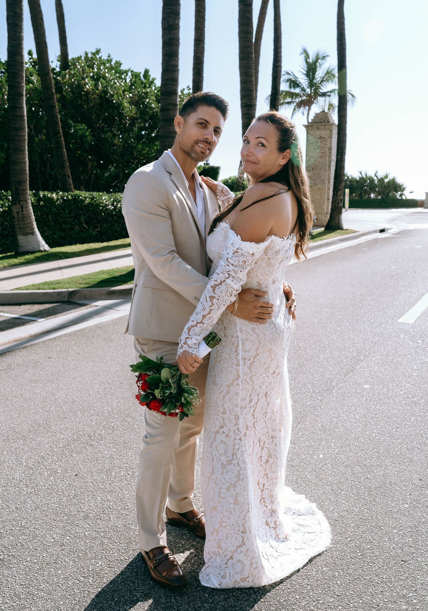 Wedding couple posing on palm-lined road, bride in lace gown holding red roses.