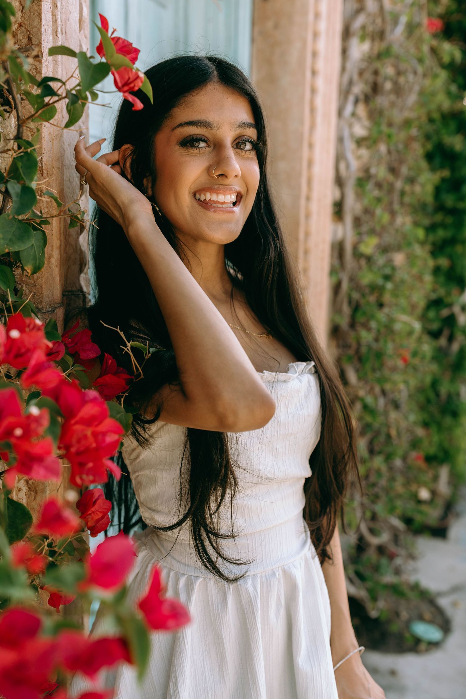 Young woman in white dress smiling among red bougainvillea flowers against a warm wall on Worth Avenue Palm beach.