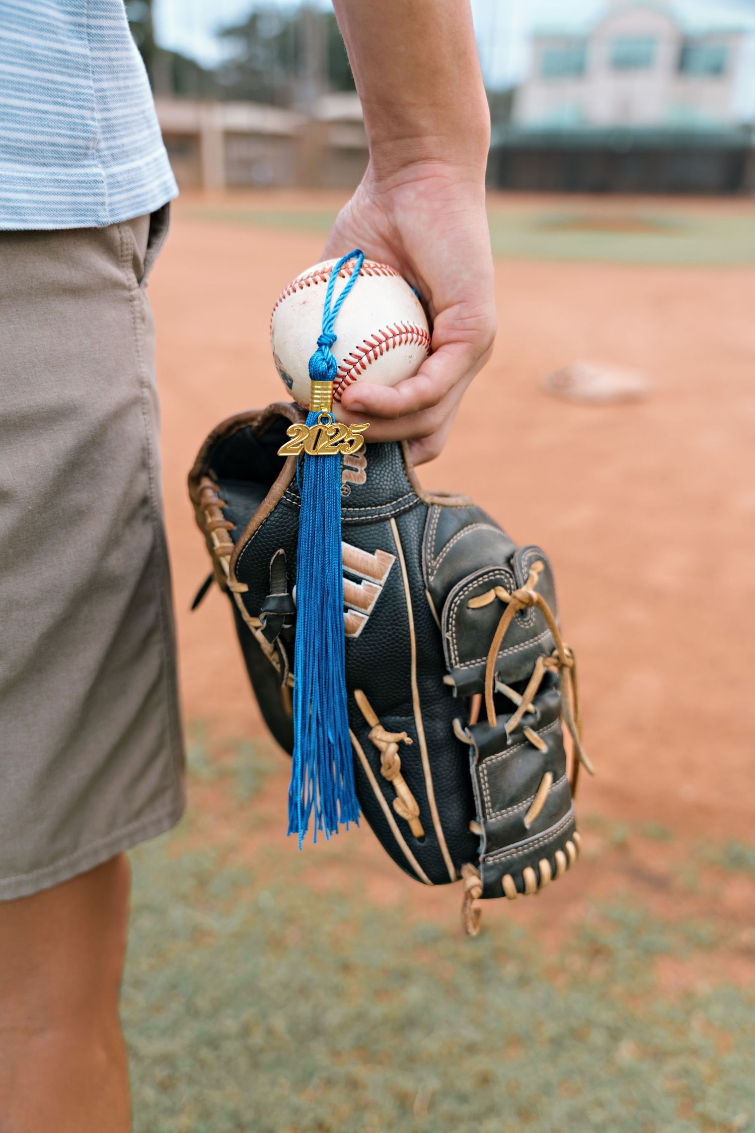 Baseball glove and ball held against a dirt field background with blue tassels hanging from the glove.