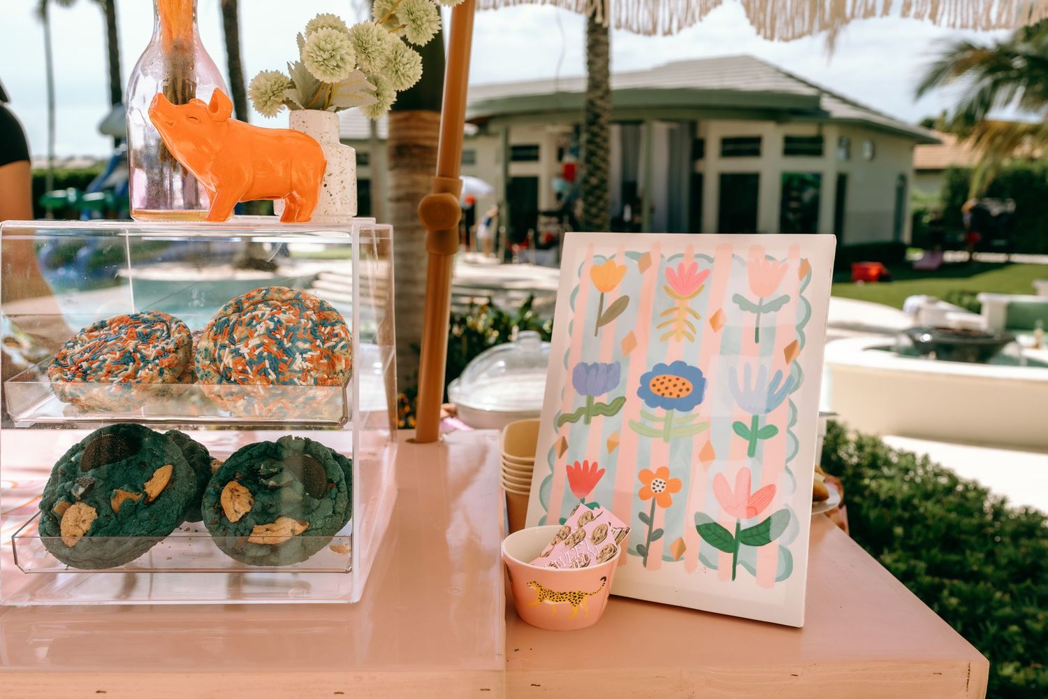 Colorful donut display case beside floral artwork at an outdoor party near a pool.