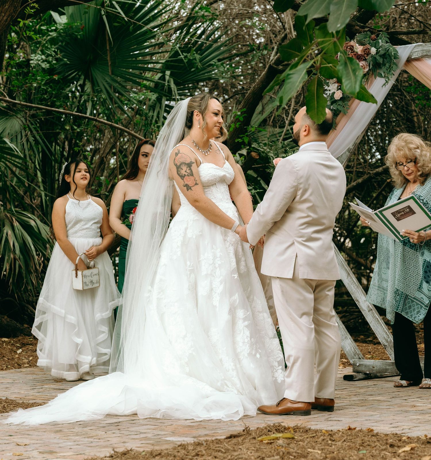 A wedding party celebrates in a lush garden setting with flowing white dresses and light-colored attire.