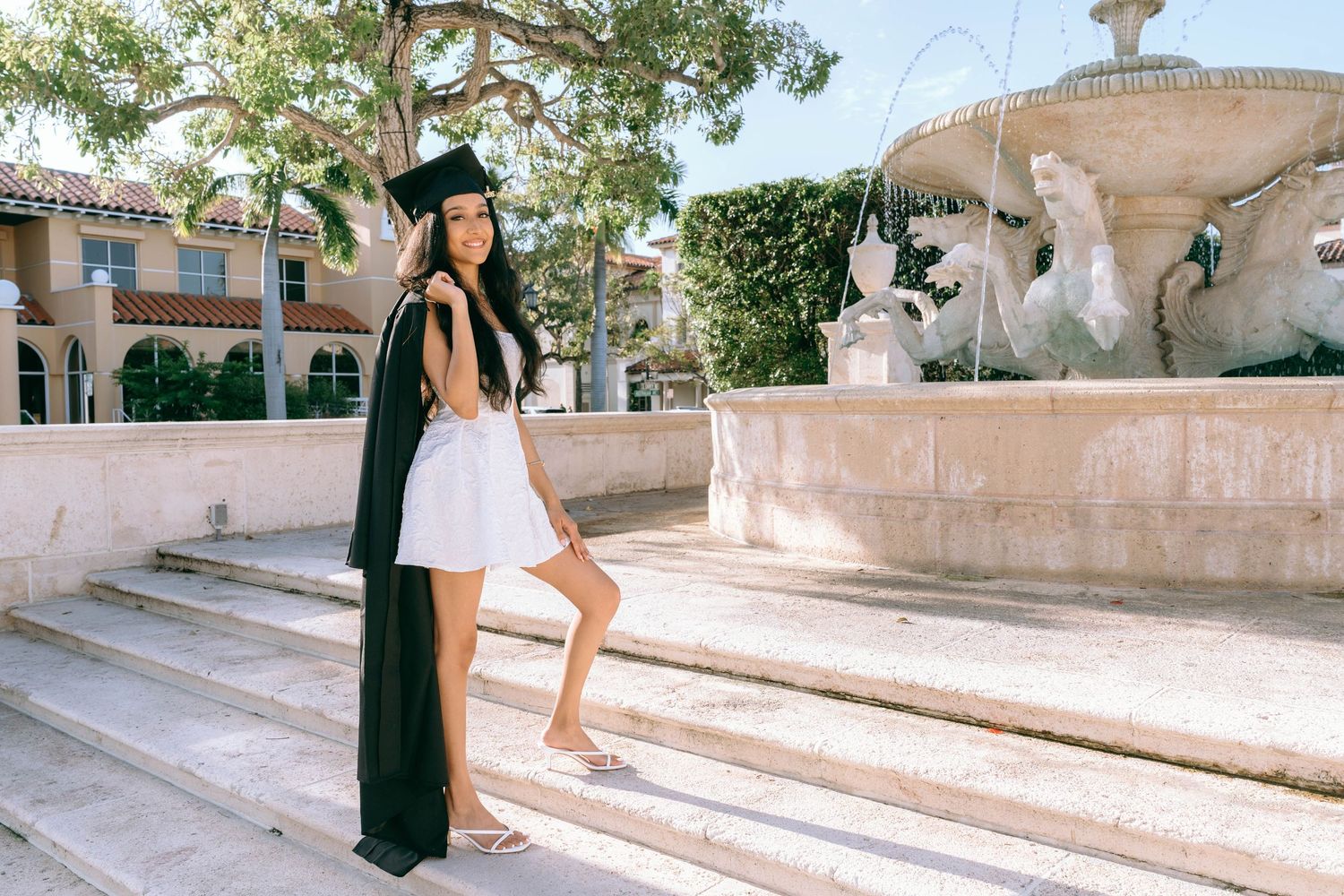 Young woman in graduation cap and gown poses by ornate fountain on sunny day on Worth Avenue Palm beach.