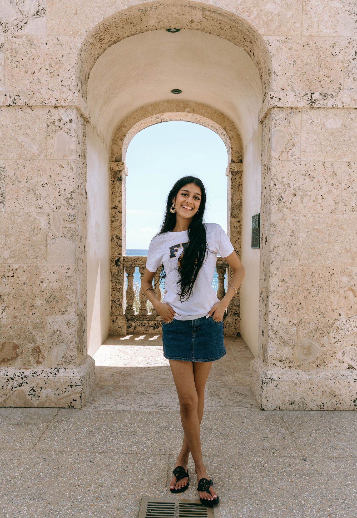 Young woman in white tee and denim skirt posing under stone arch with ocean view on Worth Avenue Palm beach.