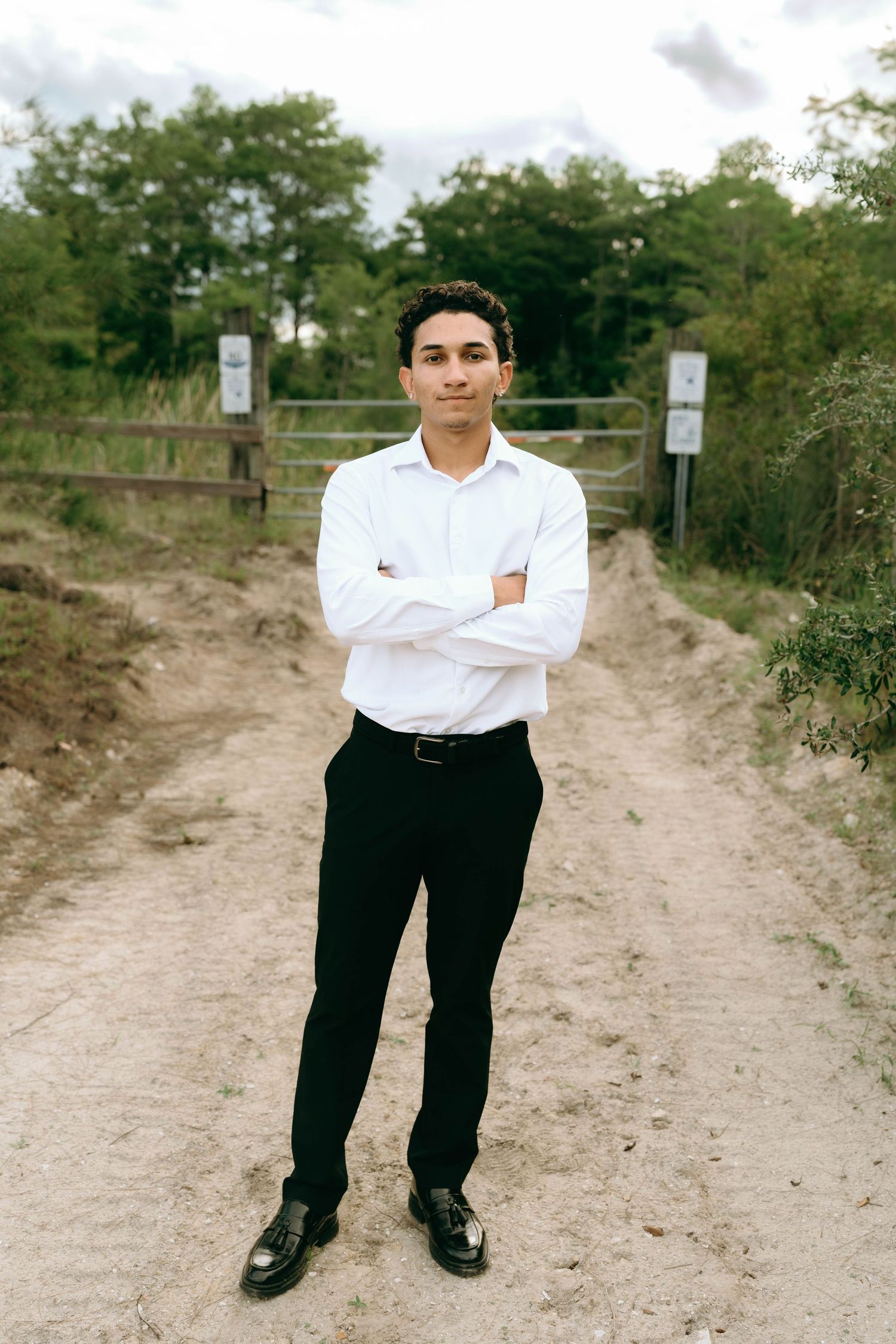 Confident young man in white shirt and black pants standing with arms crossed on a rural dirt road. Senior Photos at Colt Farms in Boynton