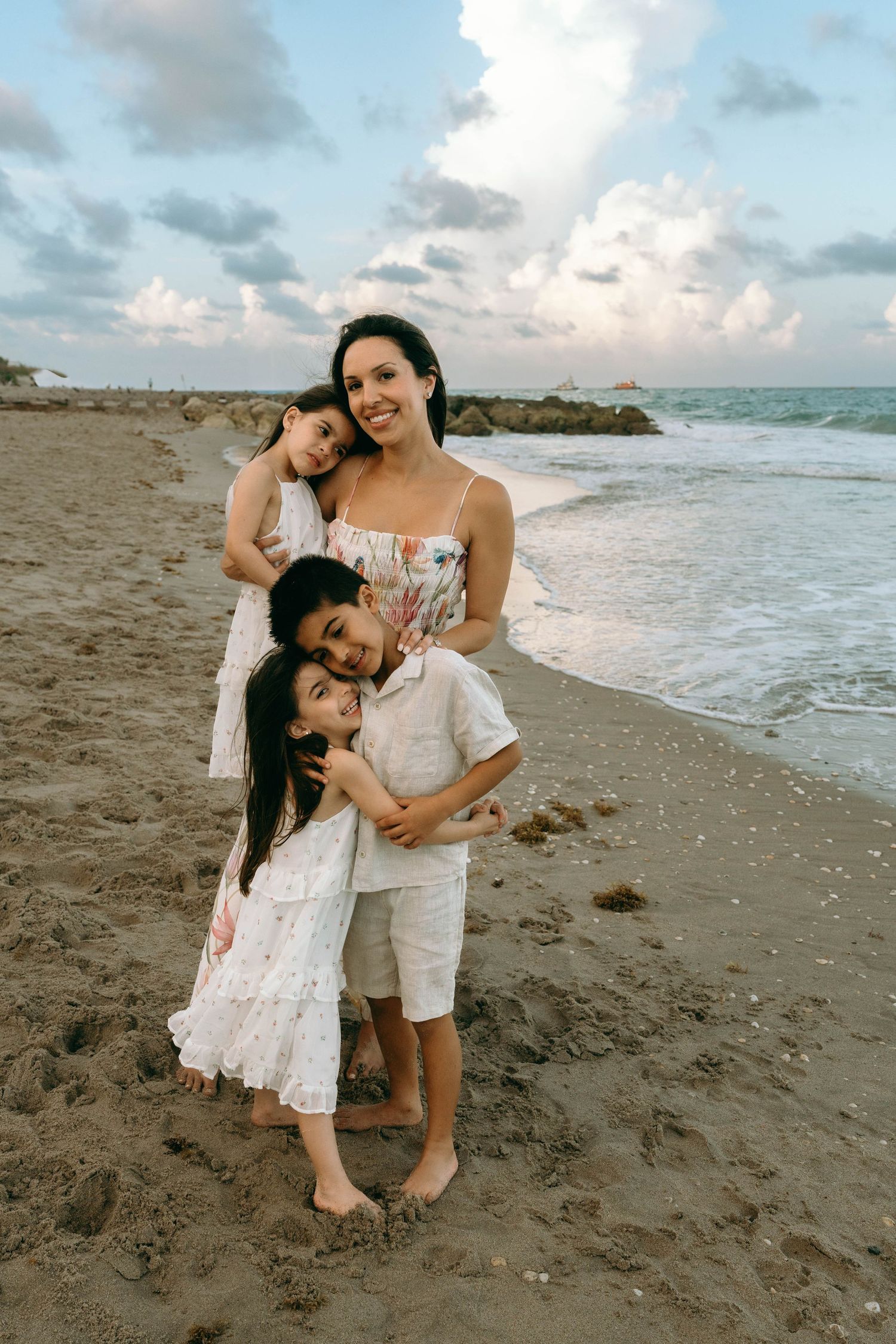 A happy mother embraces her three children on a serene beach with ocean waves at sunset. Worth Avenue, Palm Beach, FL
