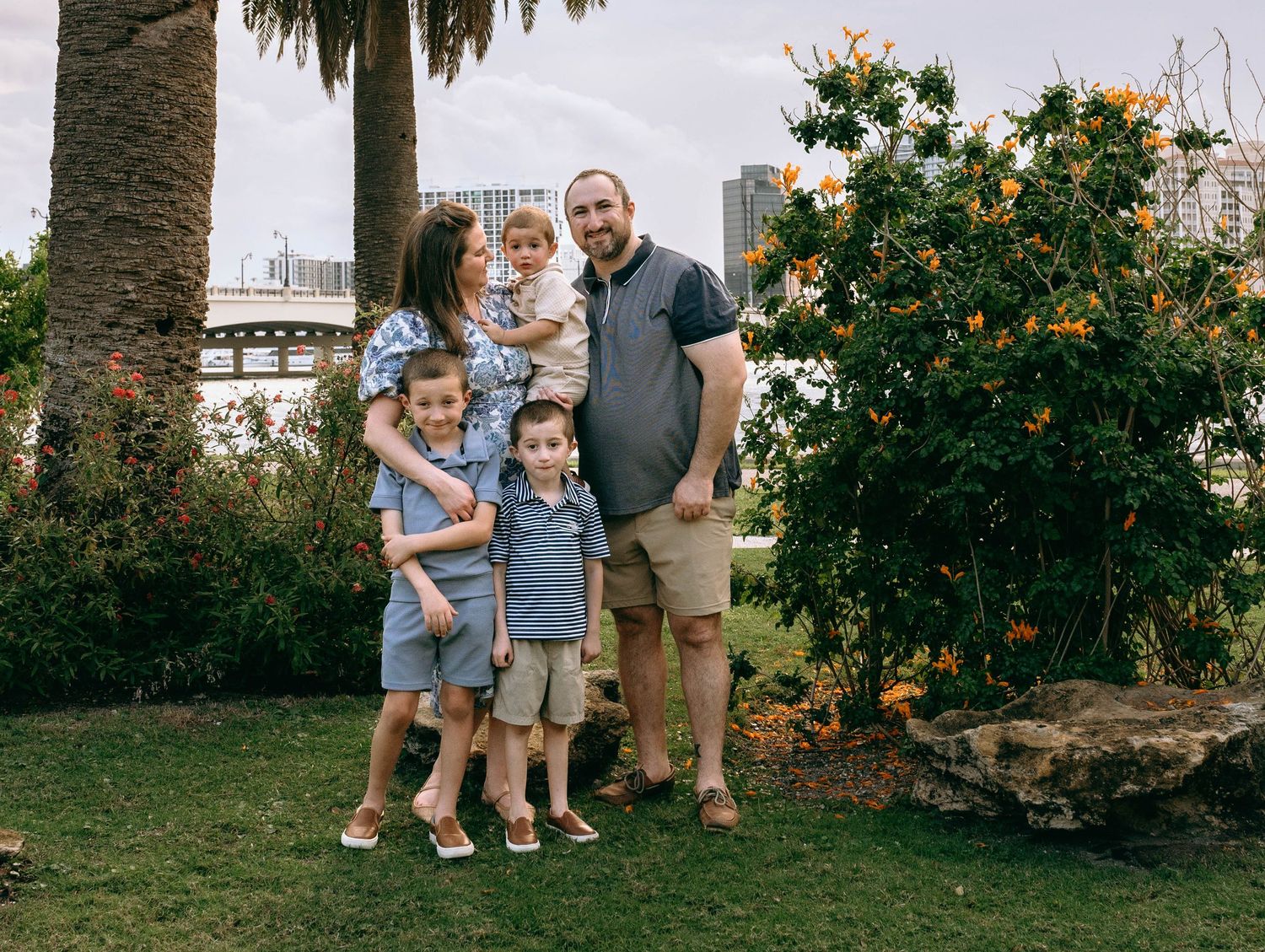 Family of five posing outdoors in a lush garden with orange flowers and city skyline in the background.  Family photos at Bradley Park Palm