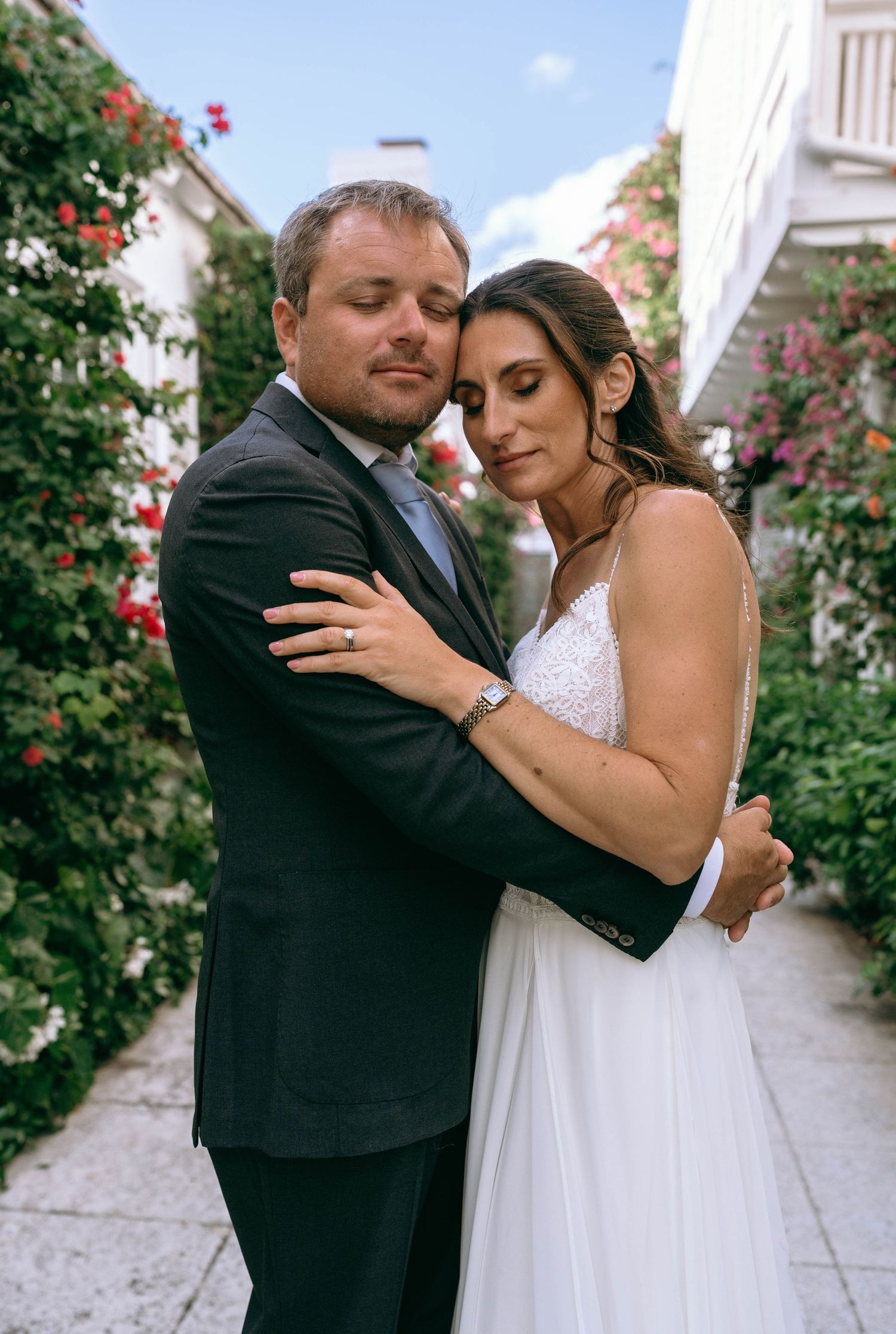 Wedding couple embracing tenderly in a floral alleyway, bride in white lace gown and groom in dark suit.