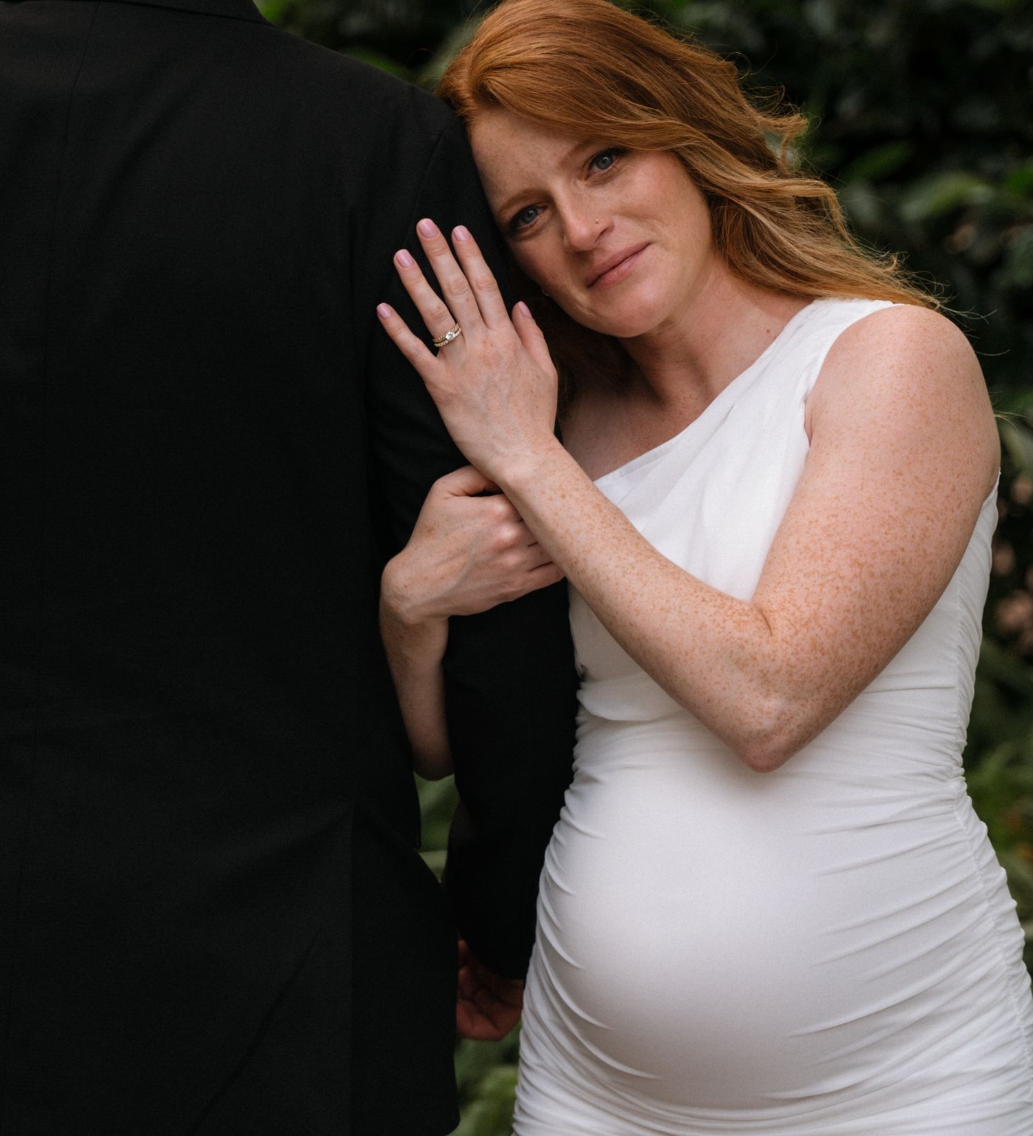 Expectant mother in a white dress poses tenderly against a dark backdrop in an outdoor maternity photo.