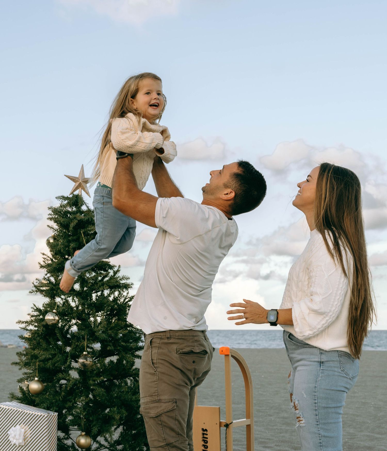 Family celebrating Christmas outdoors near the ocean, father lifting laughing daughter beside decorated tree.