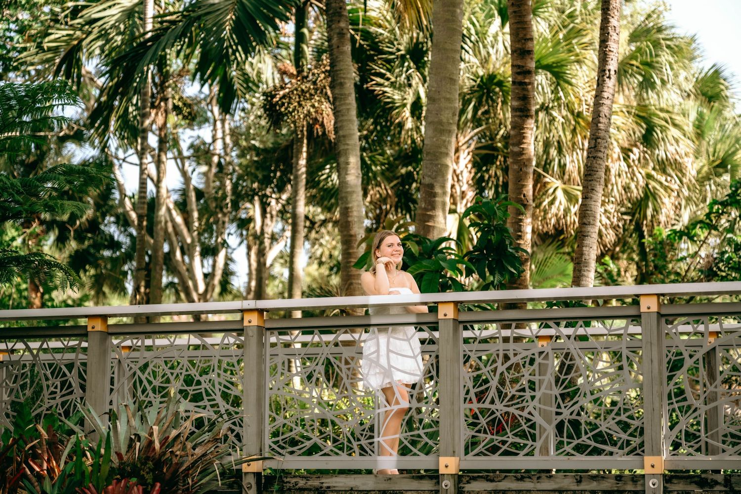 Woman in white dress standing on decorative bridge surrounded by lush tropical palm trees in a sunny garden.