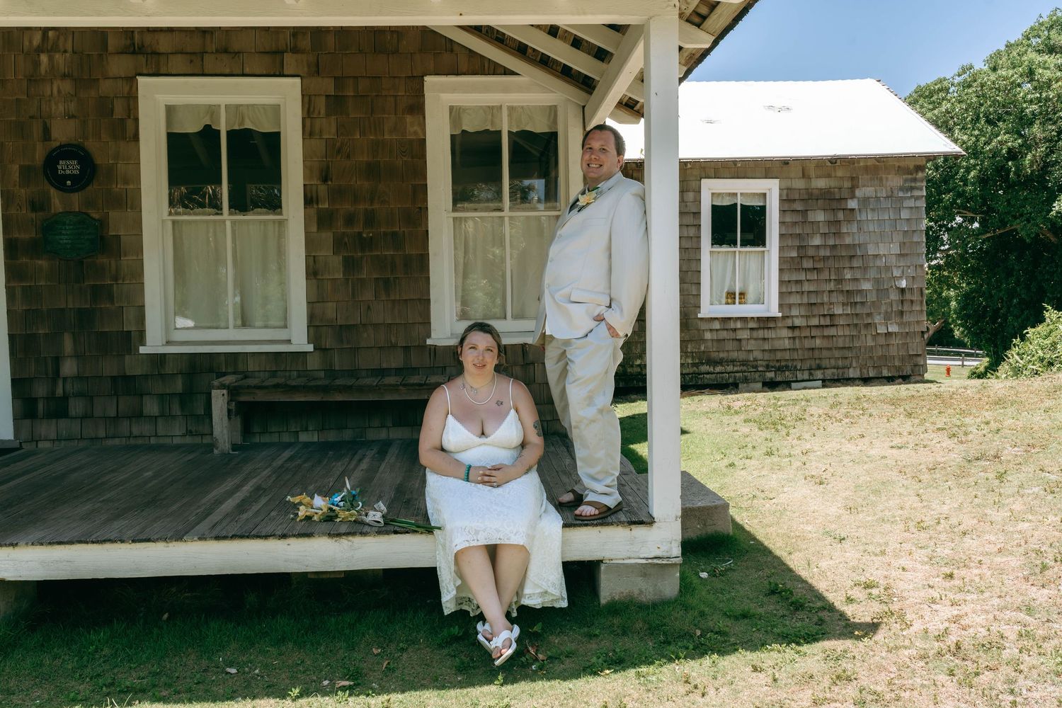 Bride and groom posing on rustic wooden porch of weathered shingle cottage on sunny wedding day.