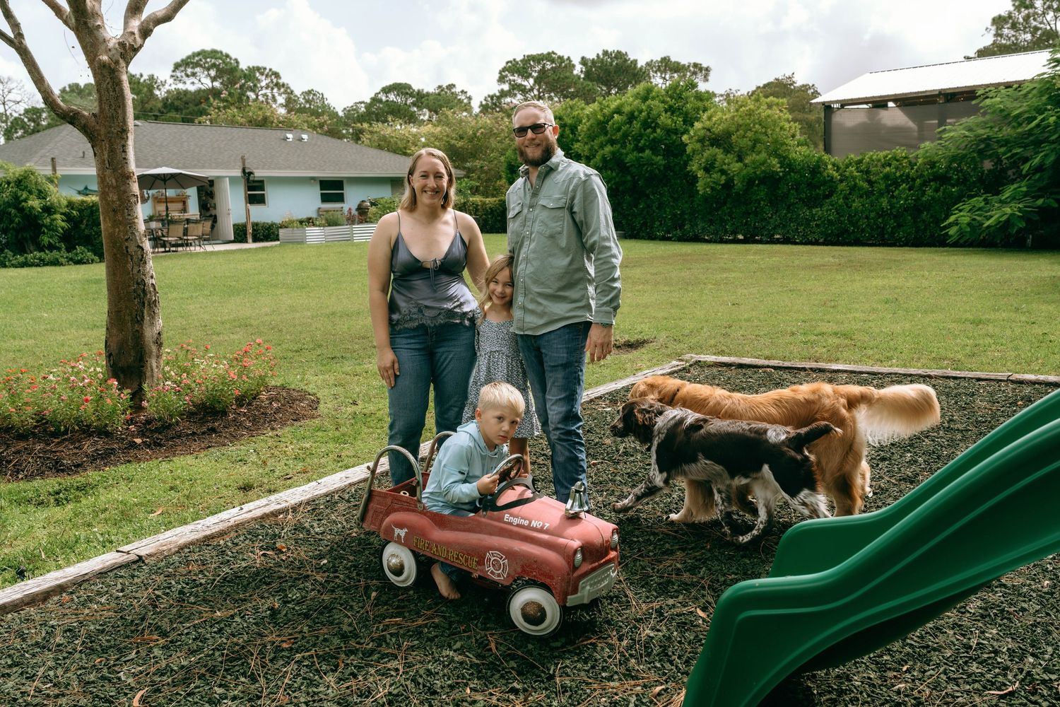 Family with two children and two dogs posing in a green backyard near a red toy car and slide.