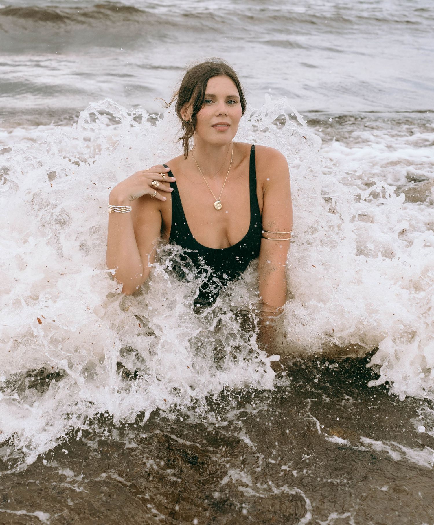 Woman in black swimsuit emerging from ocean waves on a sandy beach, wearing silver jewelry and necklace.