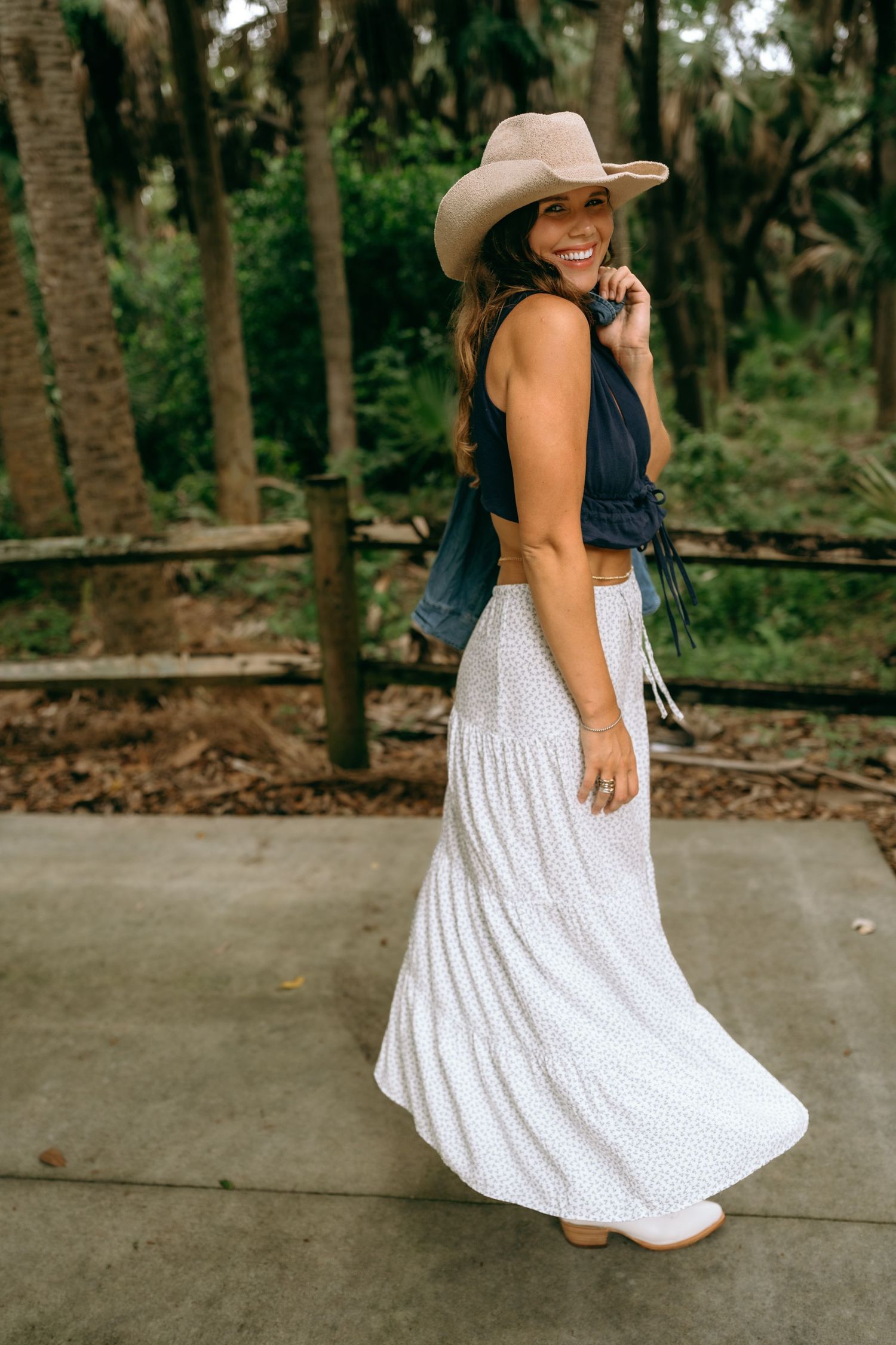 Smiling woman in navy crop top, white maxi skirt, and tan cowboy hat outdoors.
