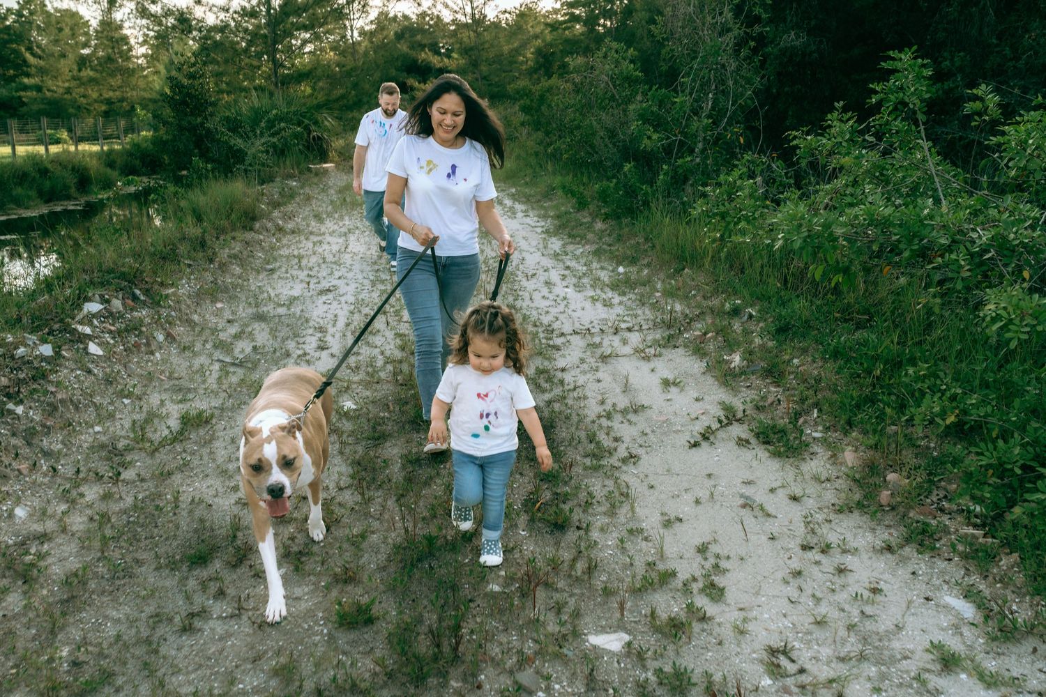 Family walking dog on leash along sandy nature trail surrounded by lush greenery.