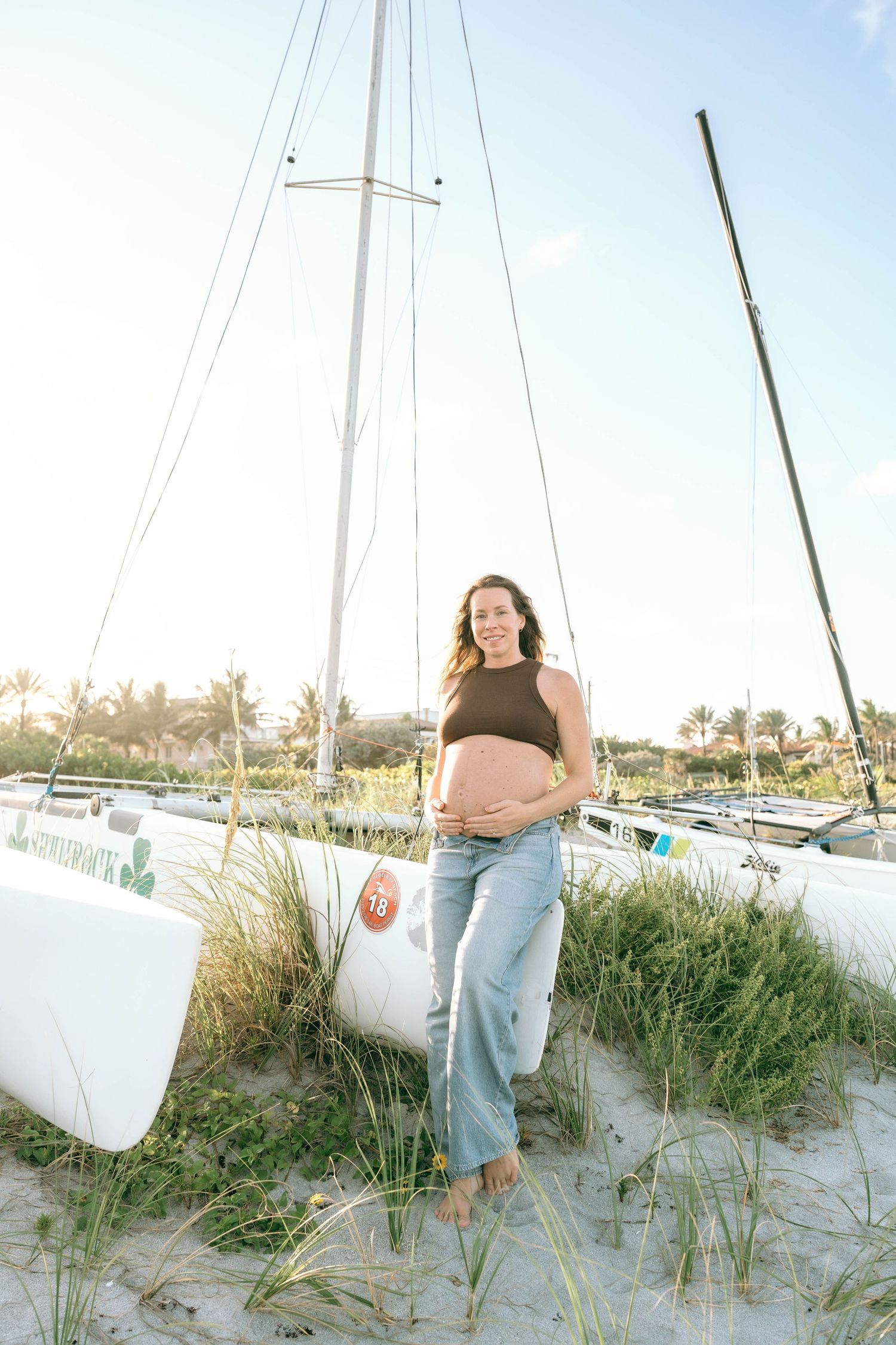 Pregnant woman in black crop top and jeans poses barefoot on sandy beach beside sailboats at golden hour.
