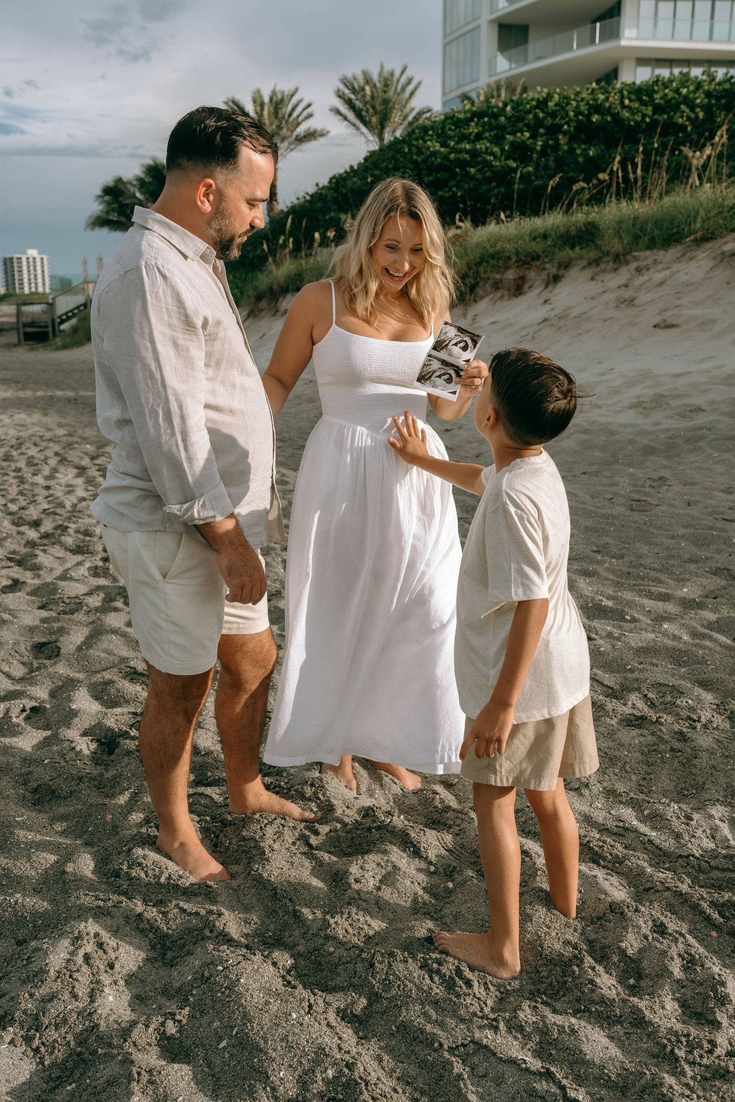 Family pregnancy announcement on beach, parents showing sonogram to young son in white outfits.