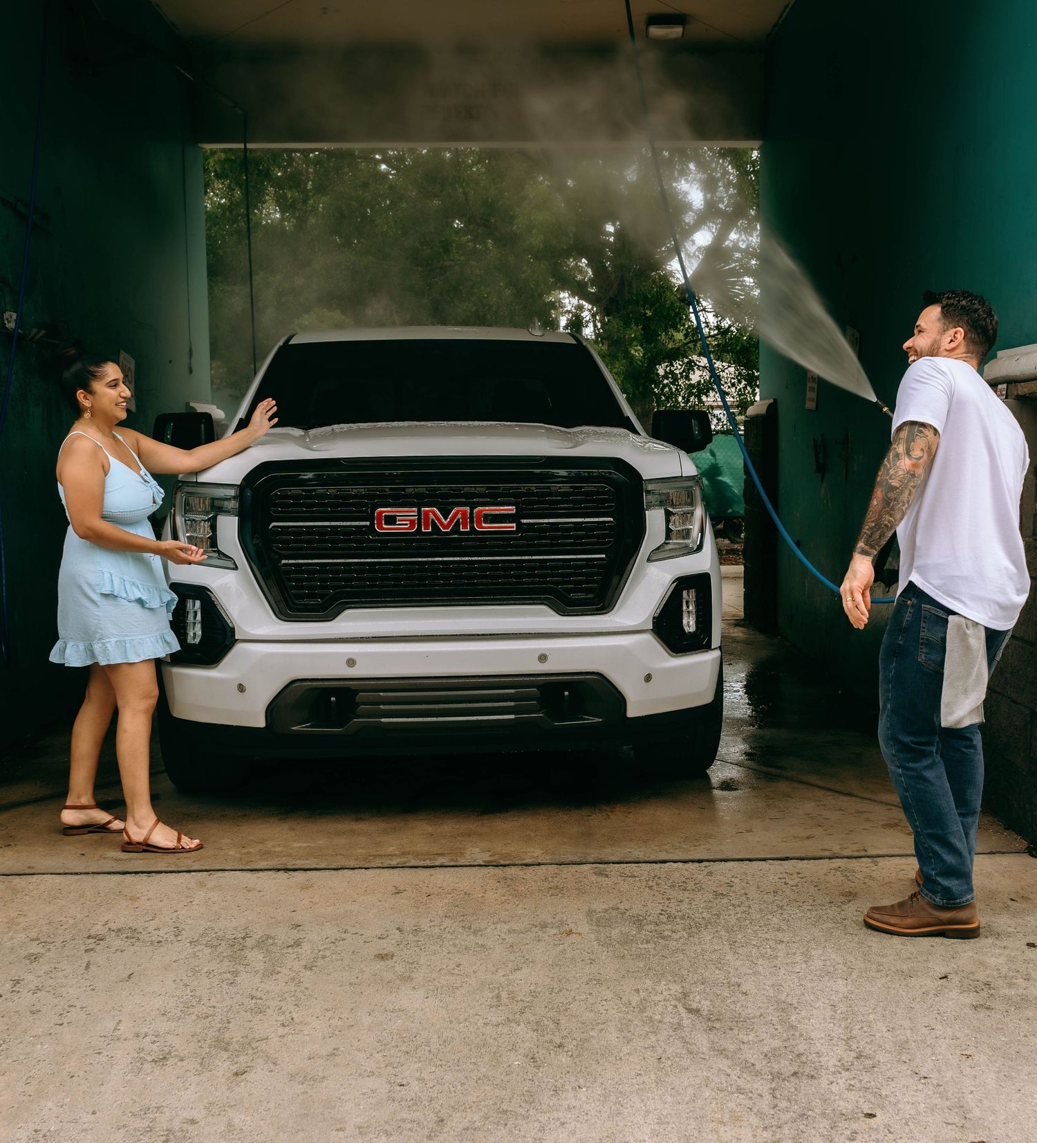 A man sprays water on a white GMC Sierra truck while a woman touches the hood during an outdoor car wash.