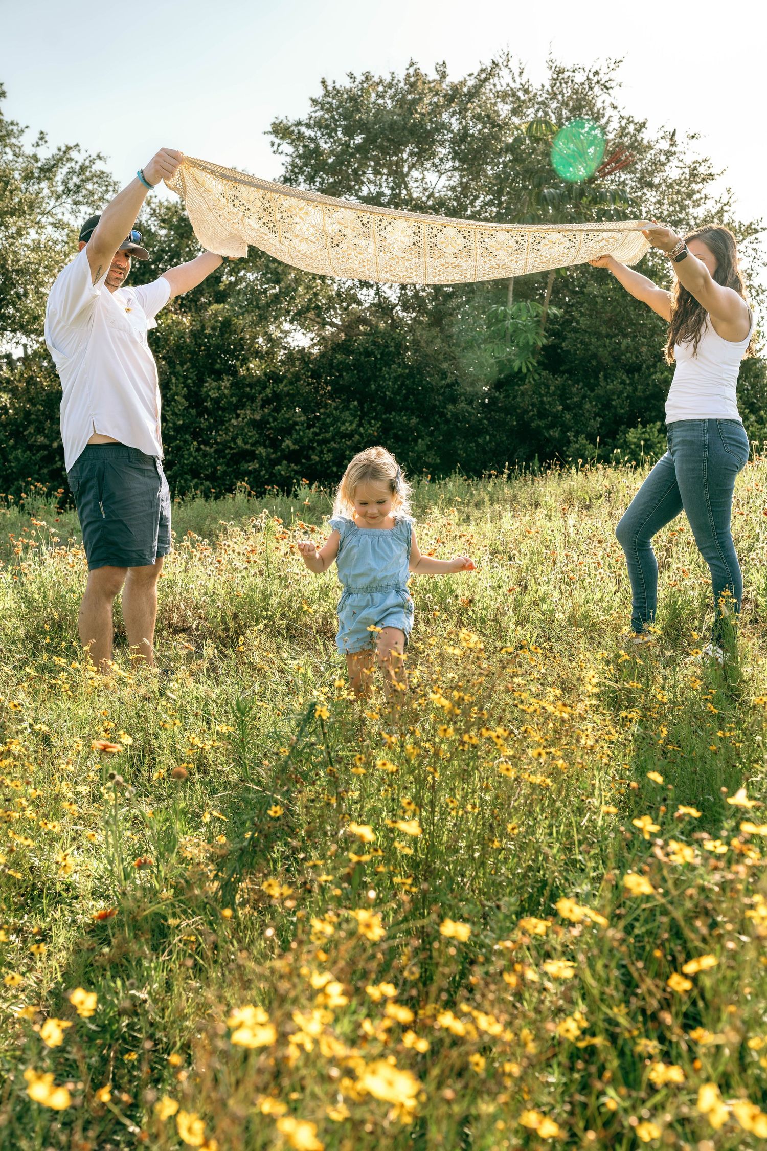 Family playing in a sunny wildflower meadow, parents holding a lace blanket over their toddler daughter.