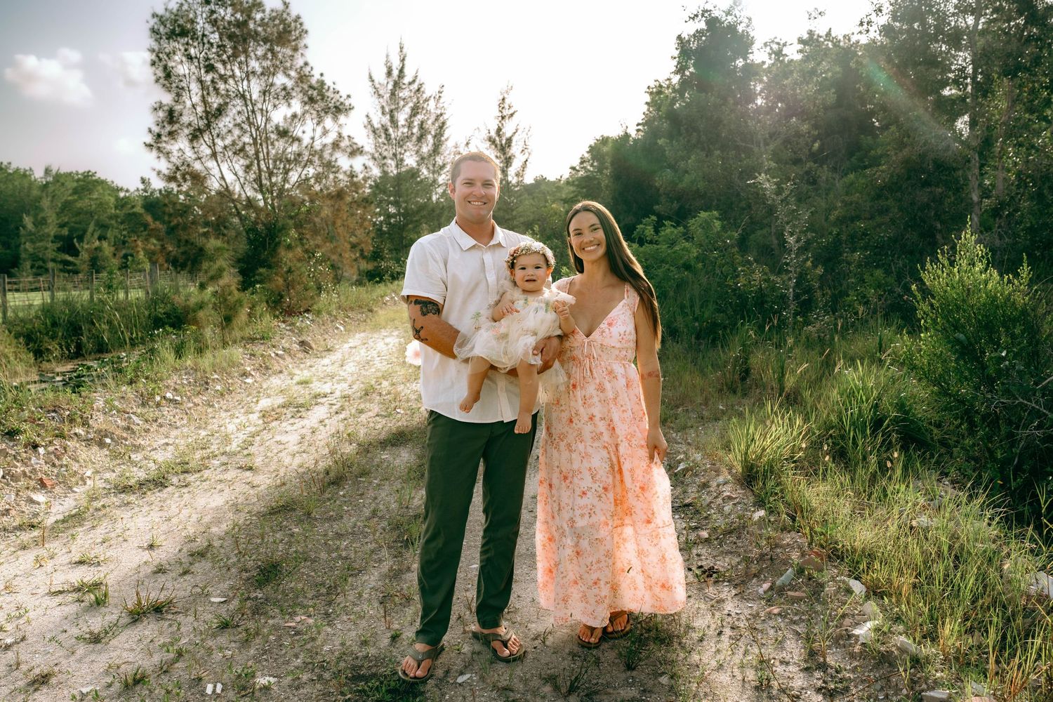 Family of three posing on a sunny dirt path surrounded by lush greenery, parents holding young child.