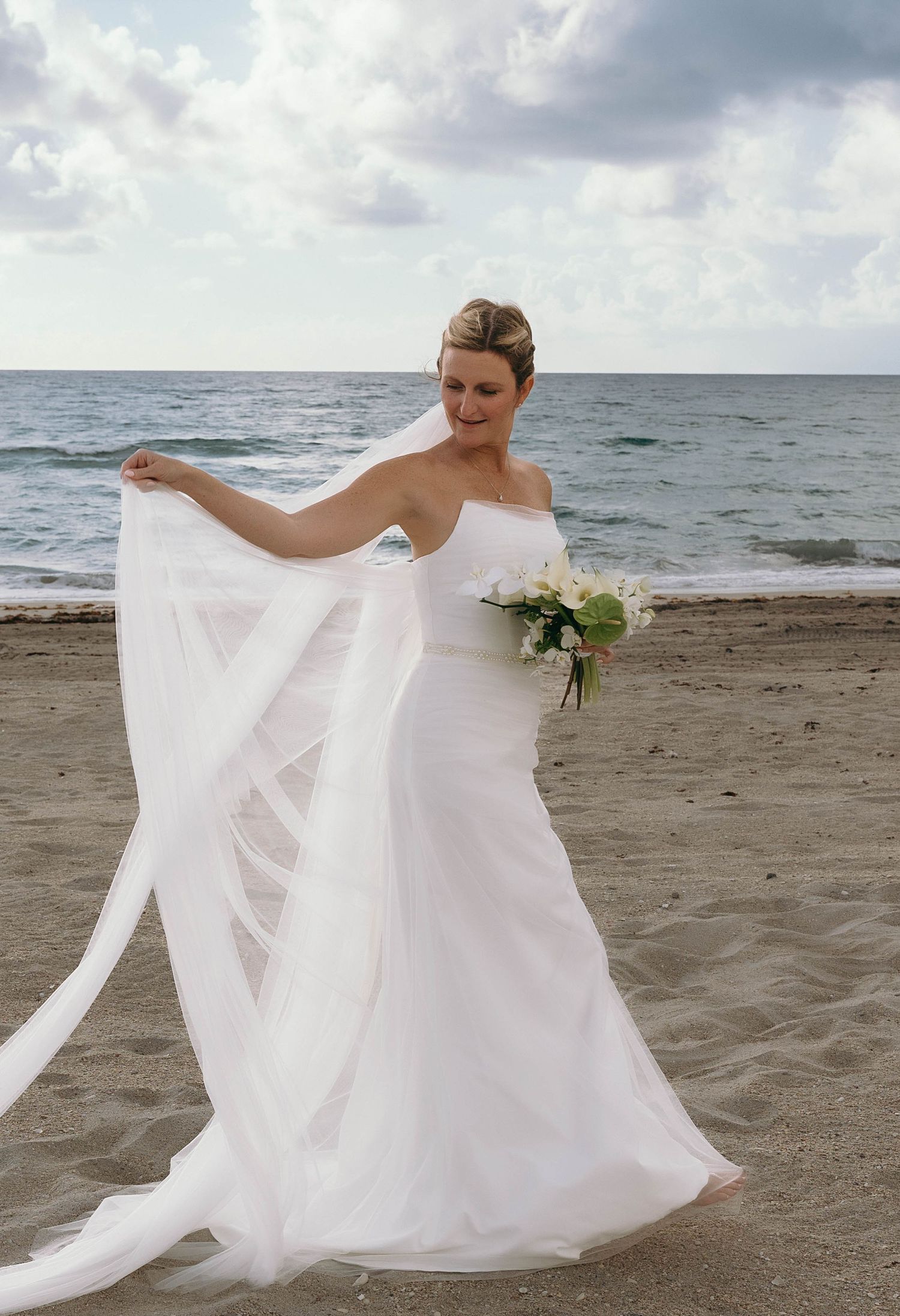 Bride twirling white wedding dress on sandy beach with ocean backdrop.