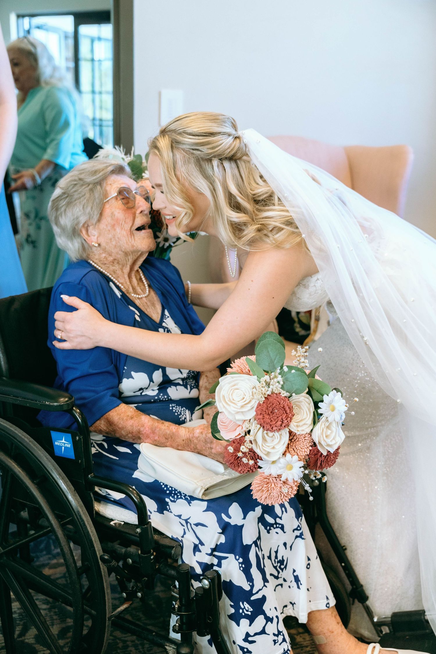 An emotional wedding moment with a bride embracing an elderly woman in a wheelchair holding a floral bouquet.