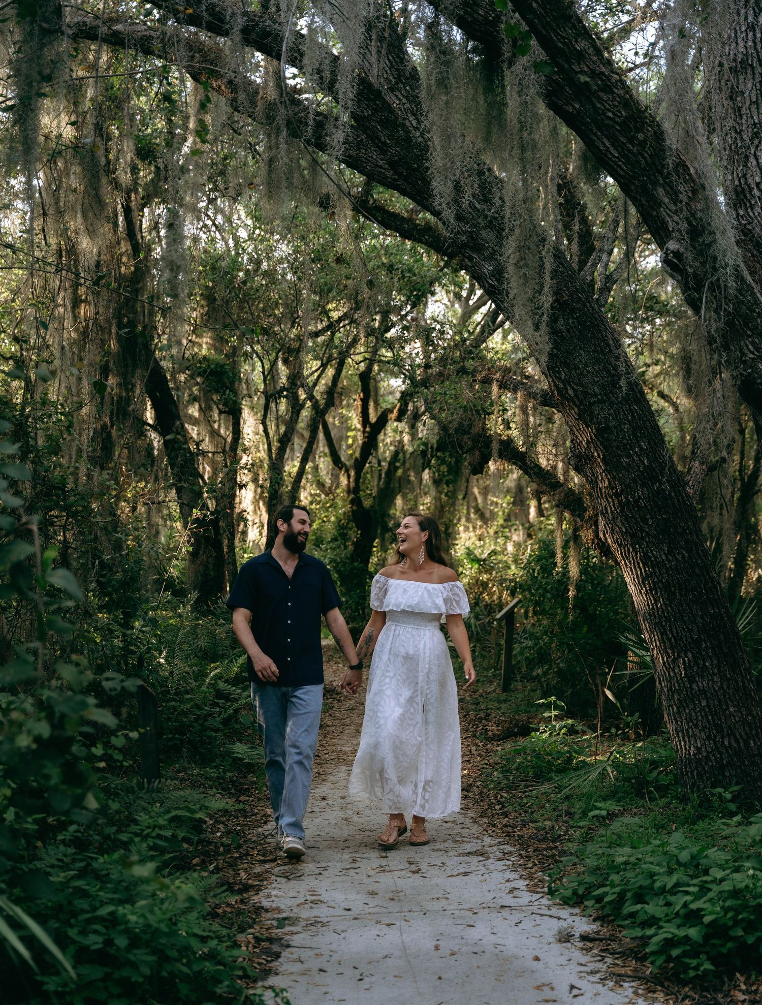 A couple walks along a winding path through a lush wooded area with Spanish moss hanging from trees.