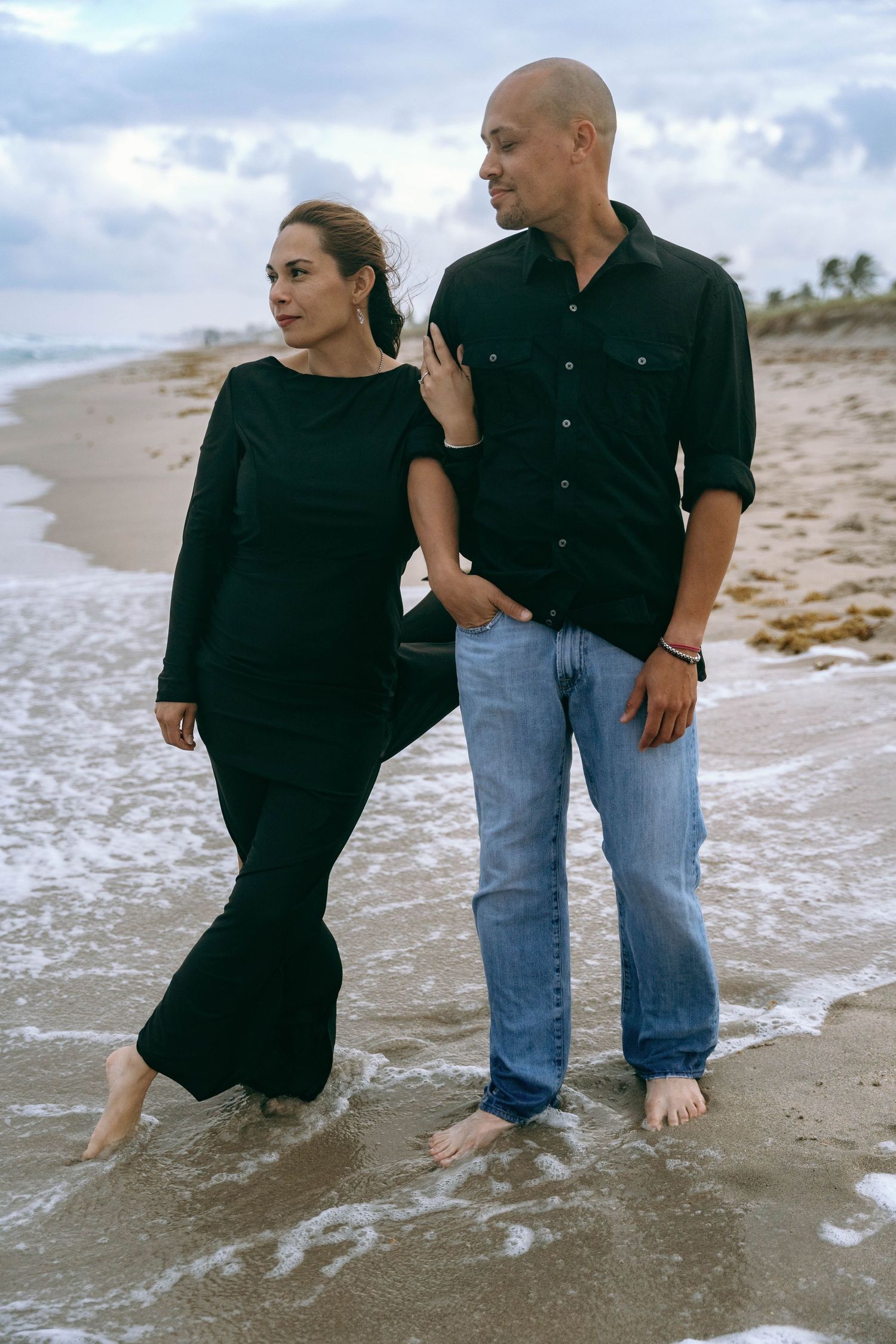 Couple walking along shoreline in black and denim outfits as waves lap at their bare feet.