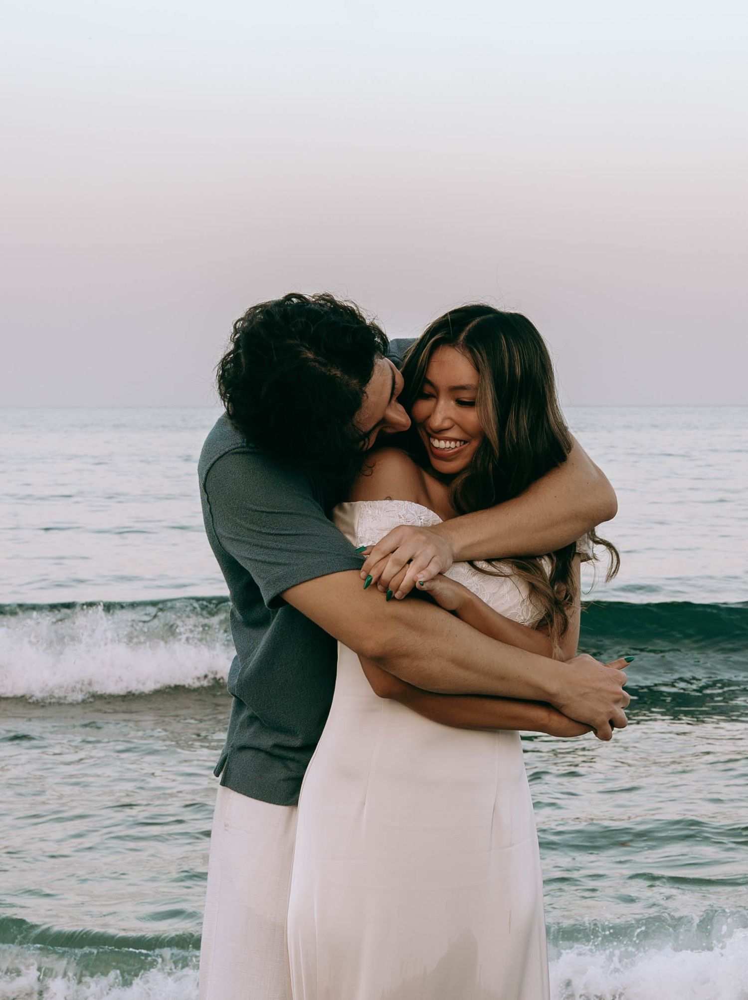 Couple embracing and laughing on a beach at sunset, woman in white dress, waves in background.