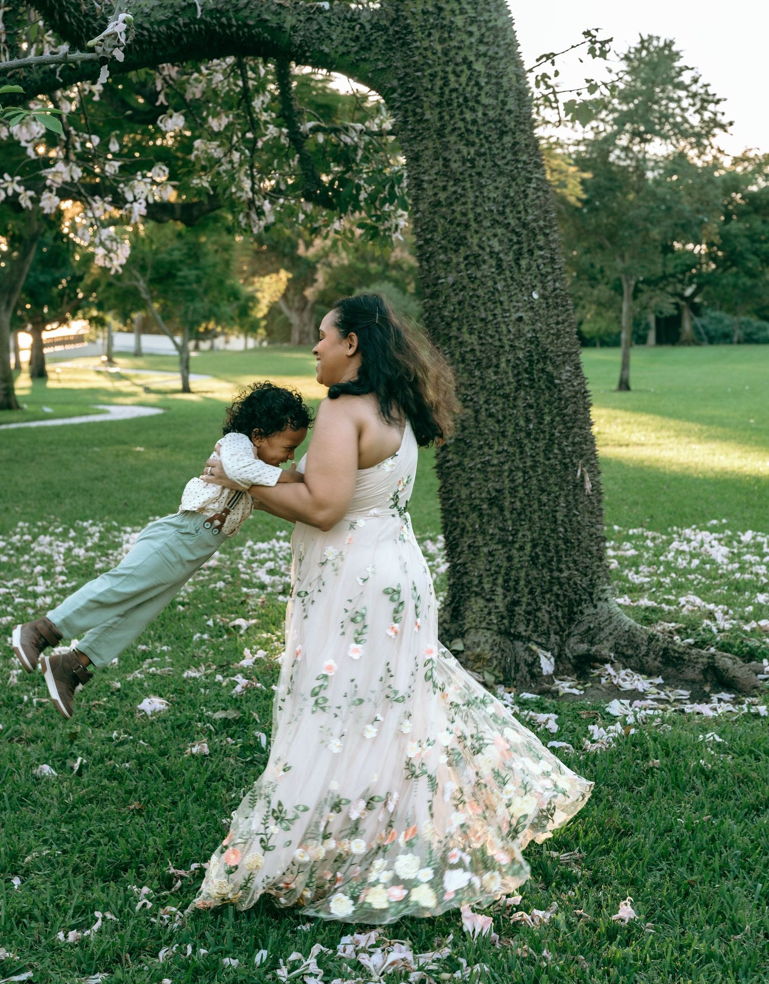 People in floral dresses dancing and twirling near a large tree trunk in a park.