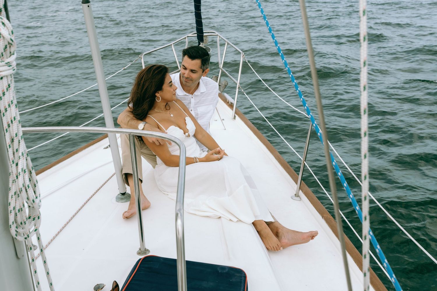 Romantic couple in white outfits sitting together on a sailboat deck on calm ocean waters.