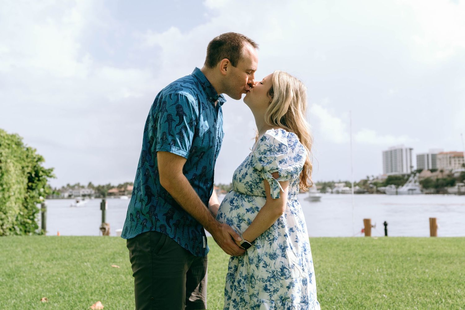 Pregnant couple sharing a romantic moment outdoors near a waterfront, man in blue floral shirt, woman in white floral dress.