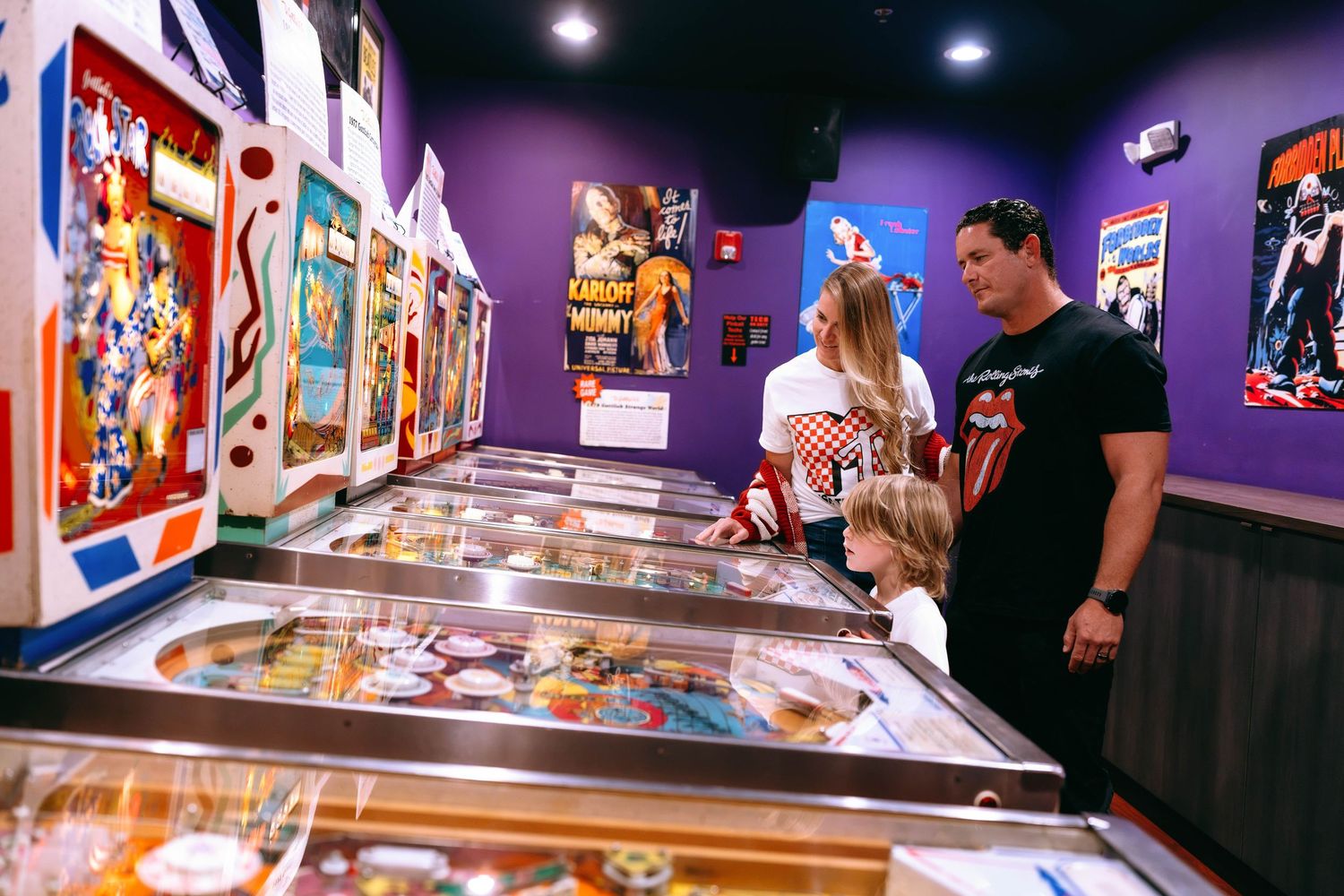 Family enjoying a vintage pinball machine arcade with colorful games lining purple walls at an entertainment venue.