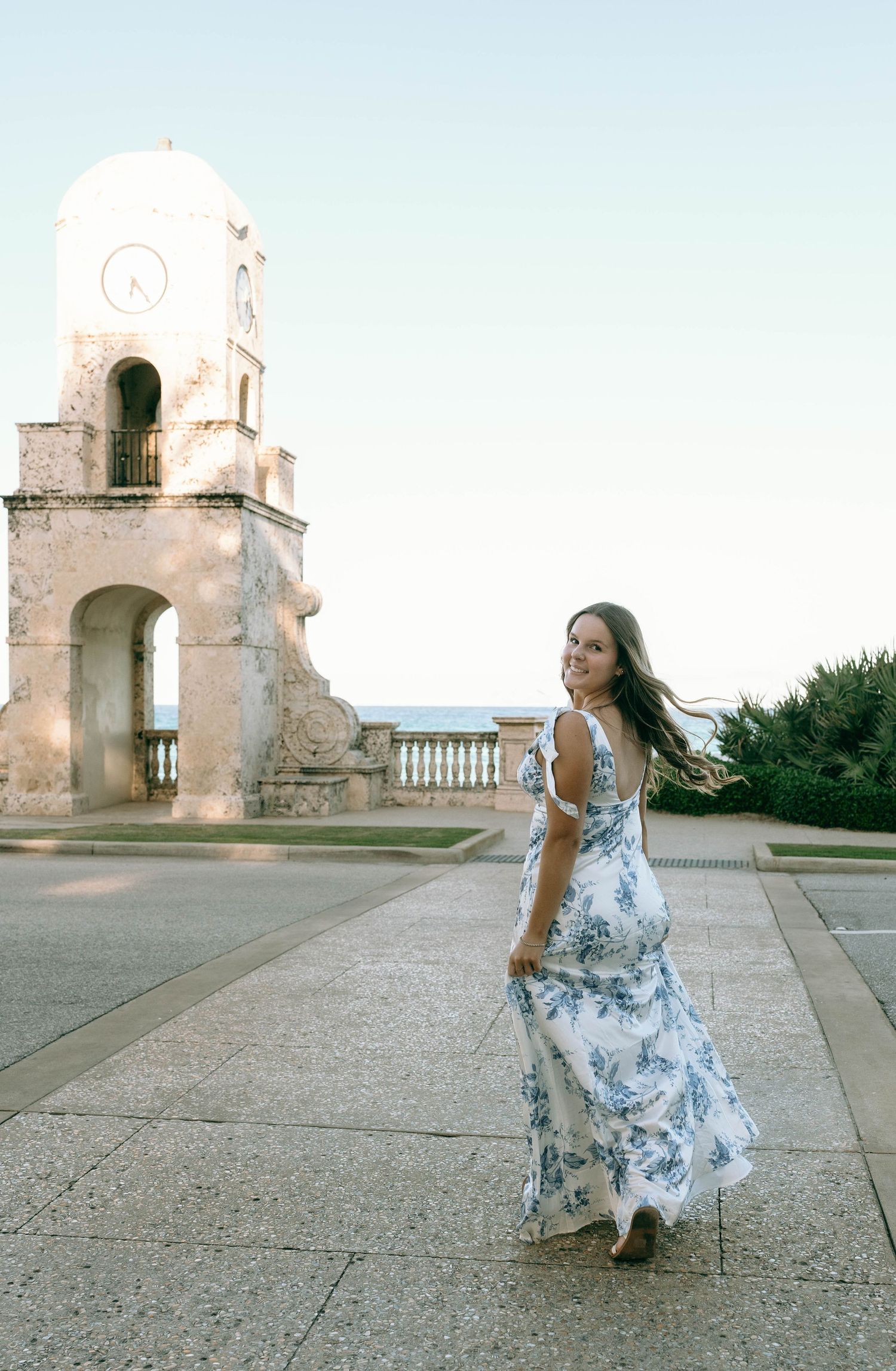Woman in flowing blue floral dress twirls near historic clock tower with ocean view backdrop.