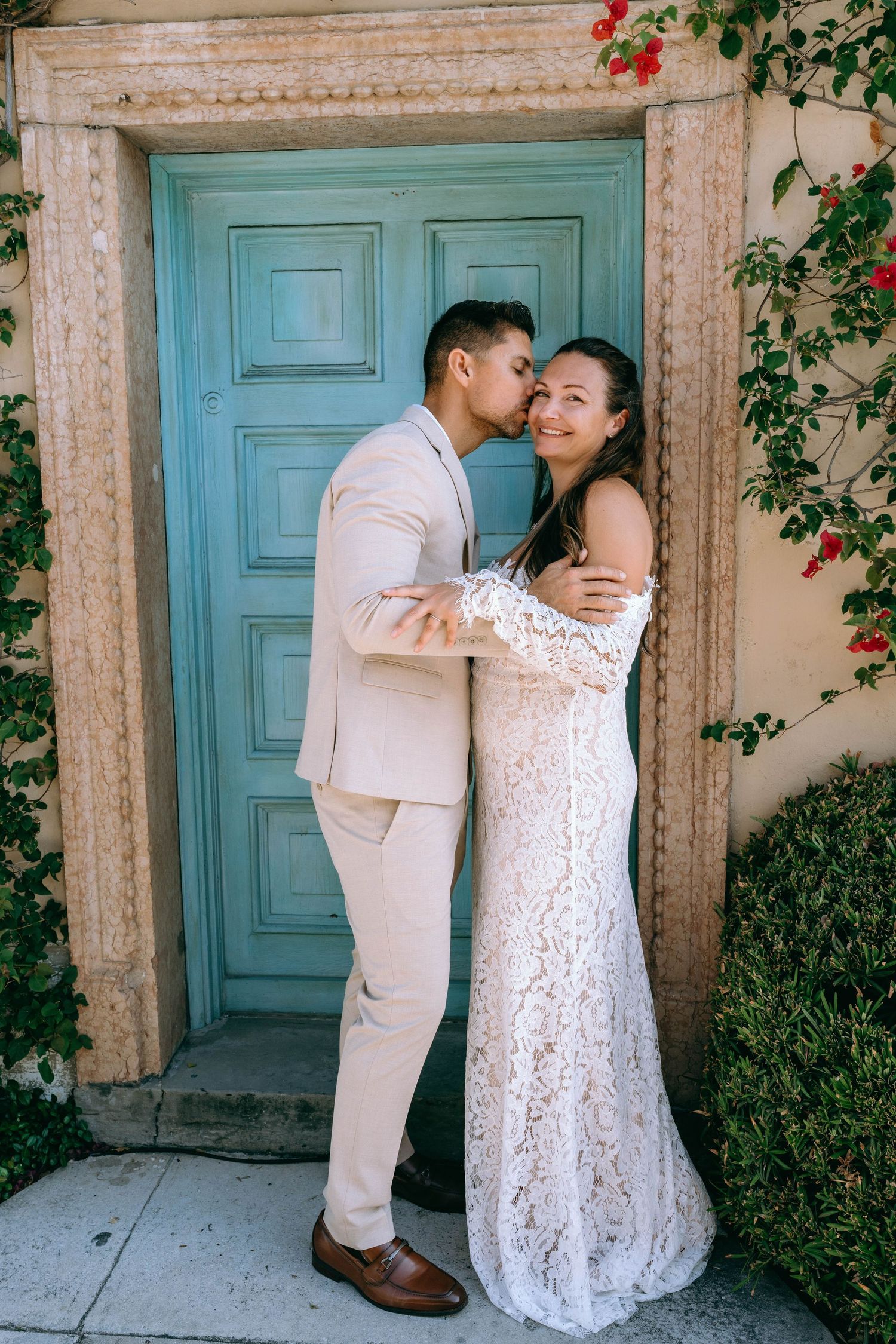 Groom kissing bride on cheek before a turquoise door adorned with red flowers, bride in white lace off-shoulder gown.