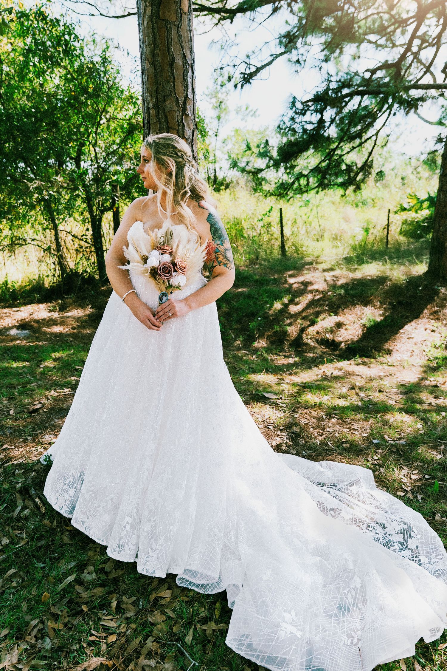 Bride in a strapless lace ball gown with a long train holds a bouquet in a sunlit forest setting.