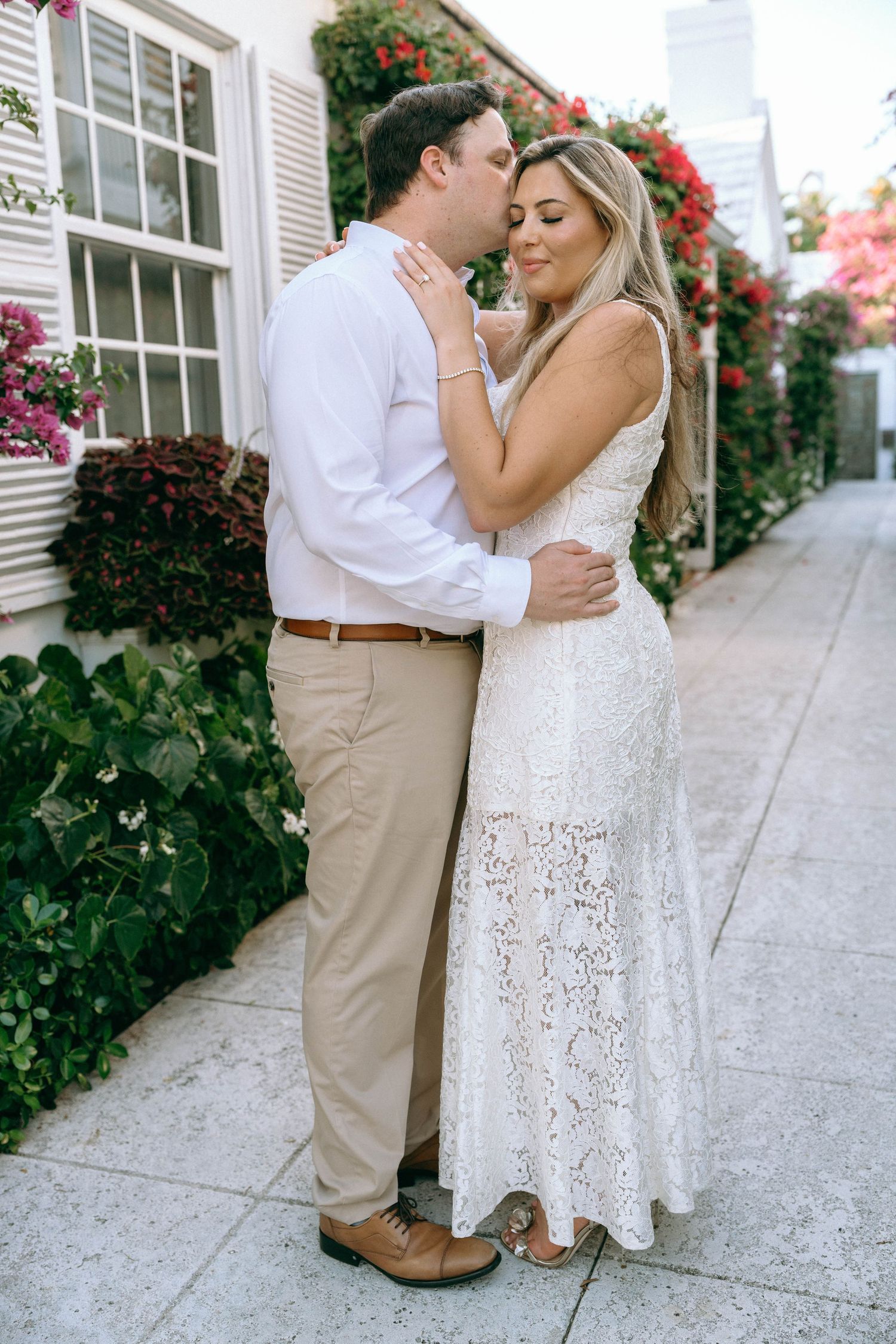 Couple embracing on a sidewalk lined with colorful flowers, man kissing womans cheek in white lace dress.