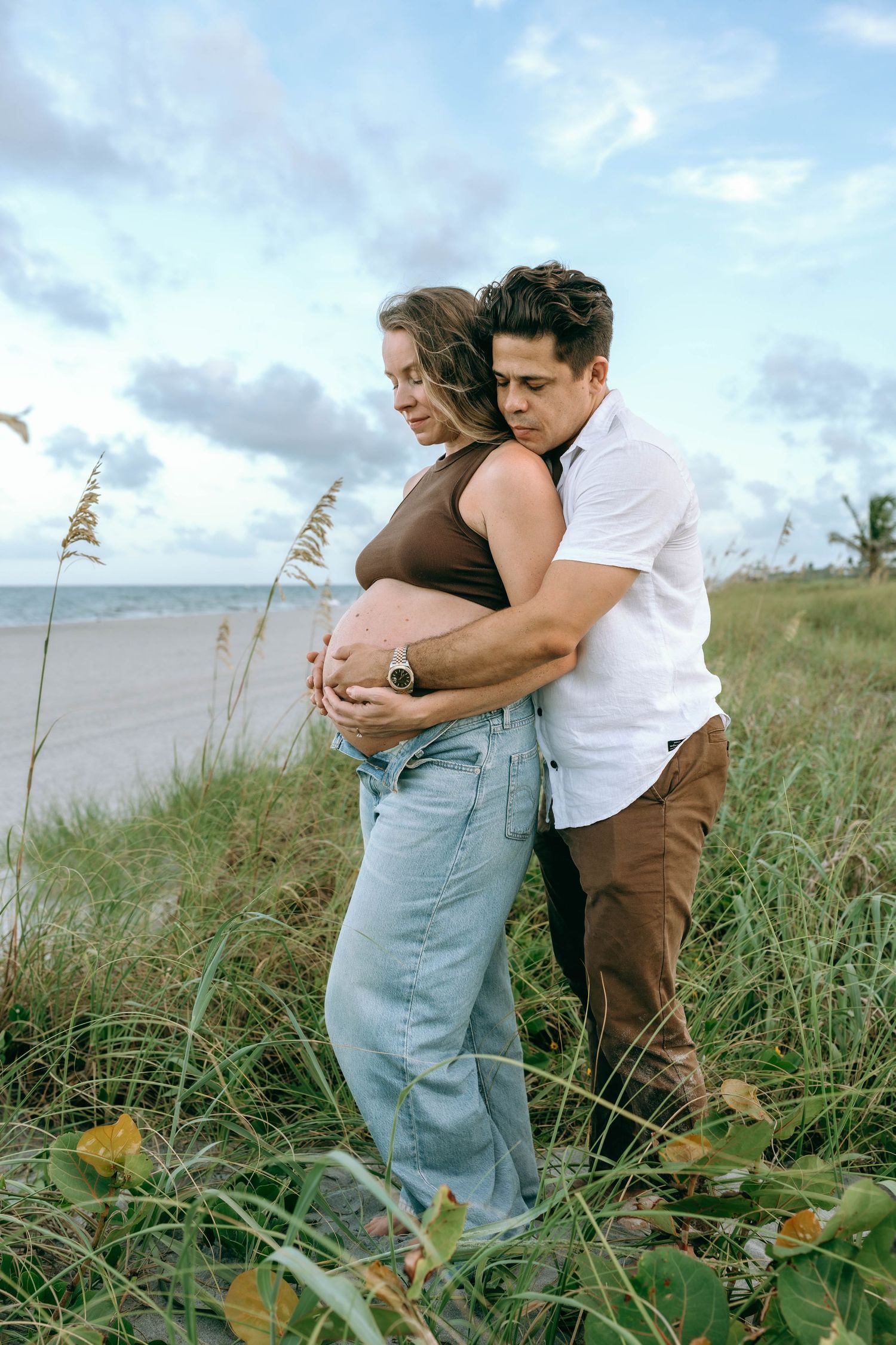 A couple embraces on a beach, the woman visibly pregnant, both gazing down tenderly at her belly near sea oats.