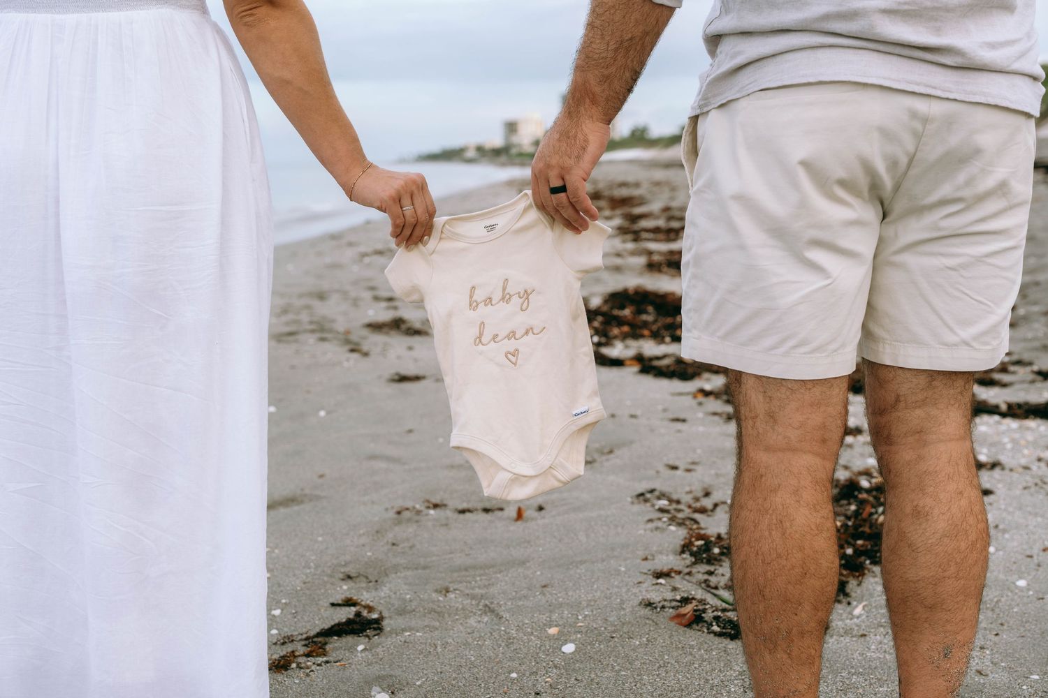 Couple holding tiny baby onesie reading baby between them on sandy beach.