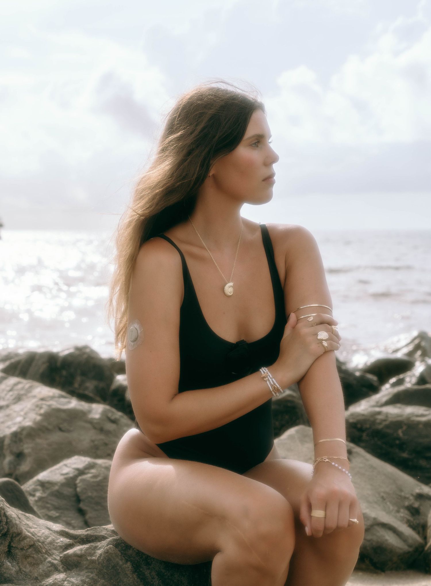 Woman in black one-piece swimsuit sitting on rocks by the ocean wearing gold jewelry and bracelets.