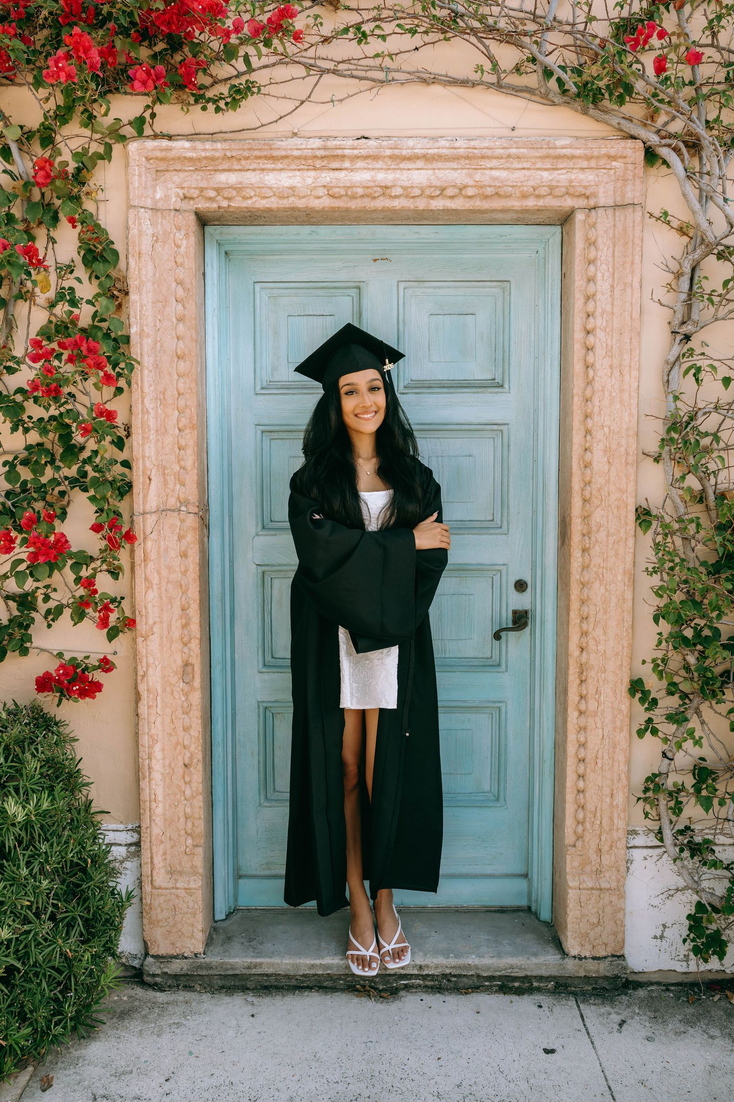 Smiling graduate in black cap and gown posing at a blue door surrounded by red flowers on Worth Avenue Palm beach.