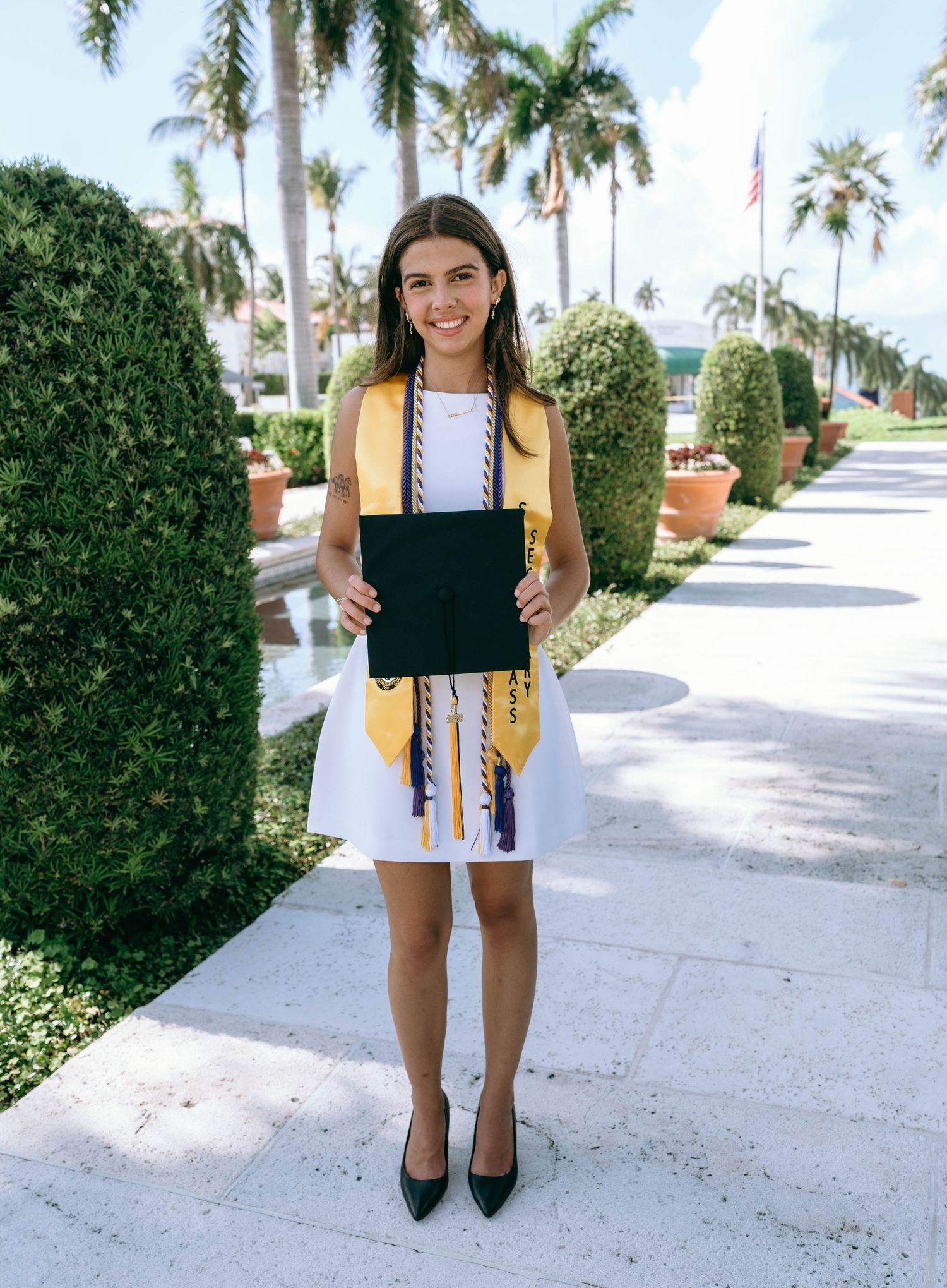 Smiling graduate in white dress with gold stole holds diploma outdoors among palm trees on a sunny day on Worth Avenue Palm beach.