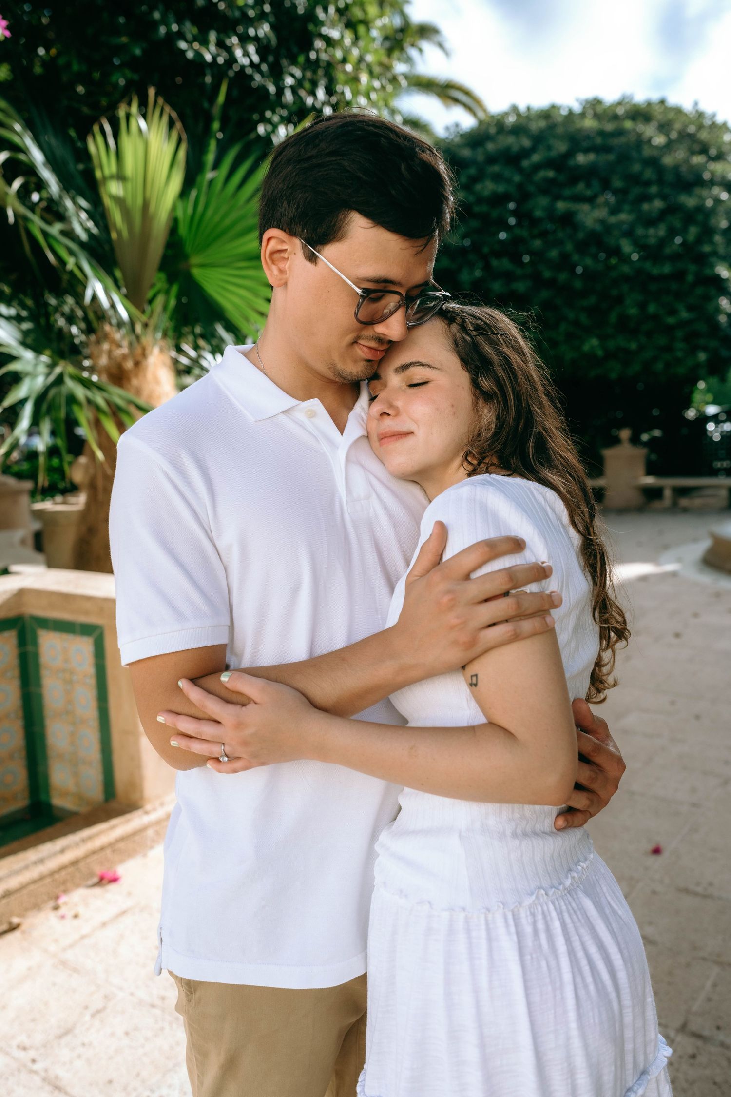 A loving couple embraces tenderly outdoors, both wearing white, surrounded by lush tropical greenery. Engagement photos on Worth Avenue