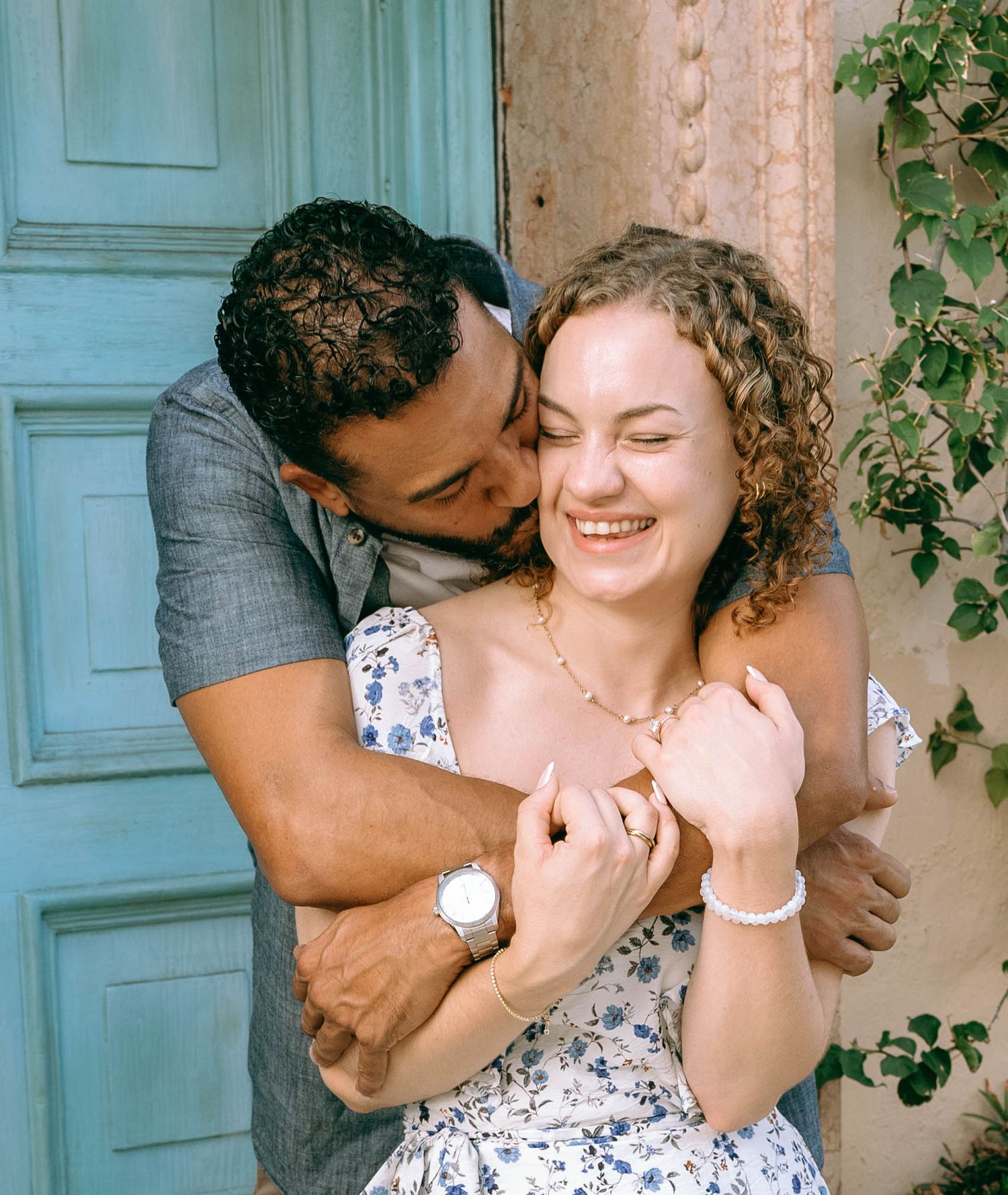 A happy couple embraces near a teal door, man kissing womans cheek as she smiles, wearing summer outfits outdoors. Engagement photos