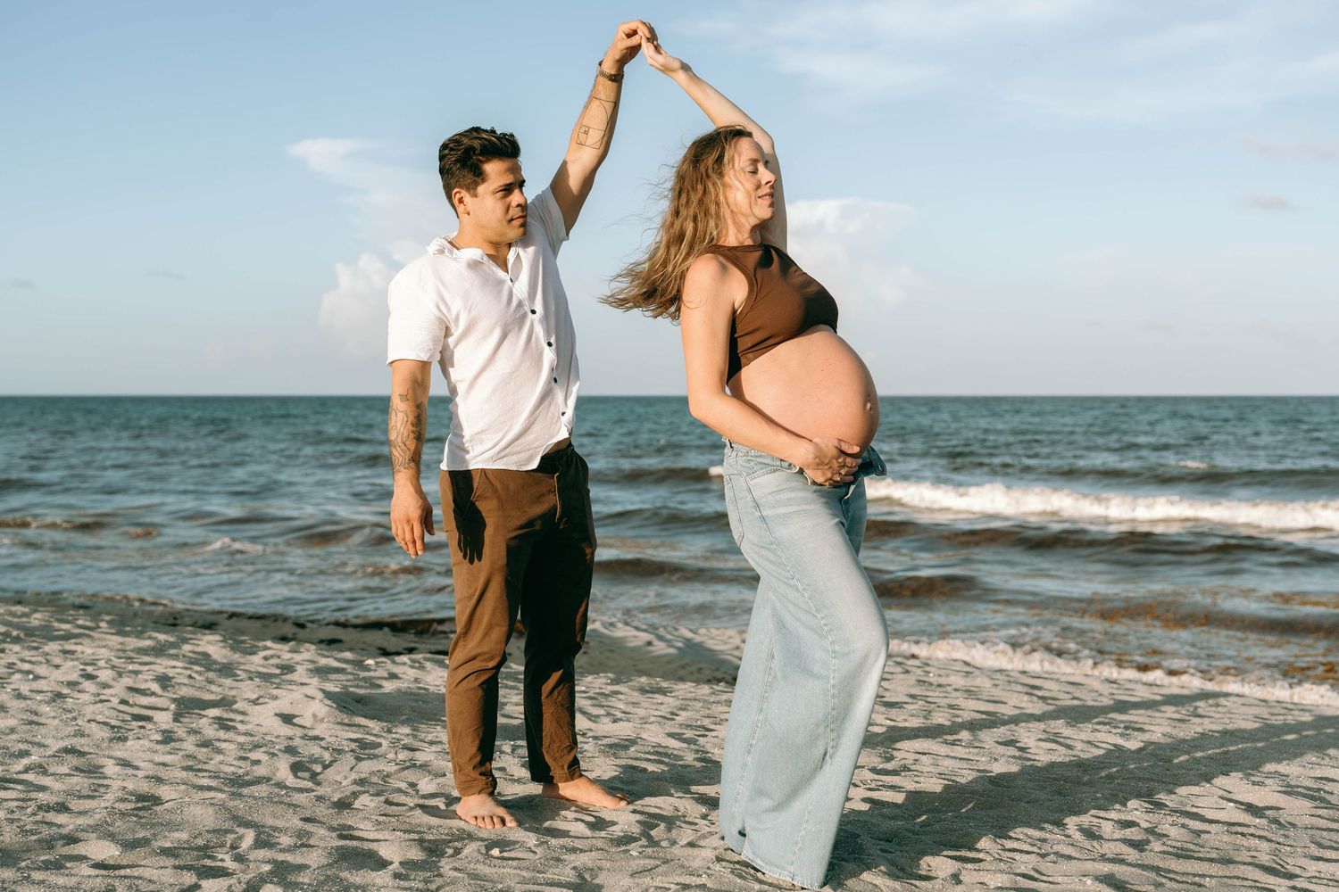 Pregnant couple dancing on beach at sunset, man spinning woman with baby bump near ocean waves.