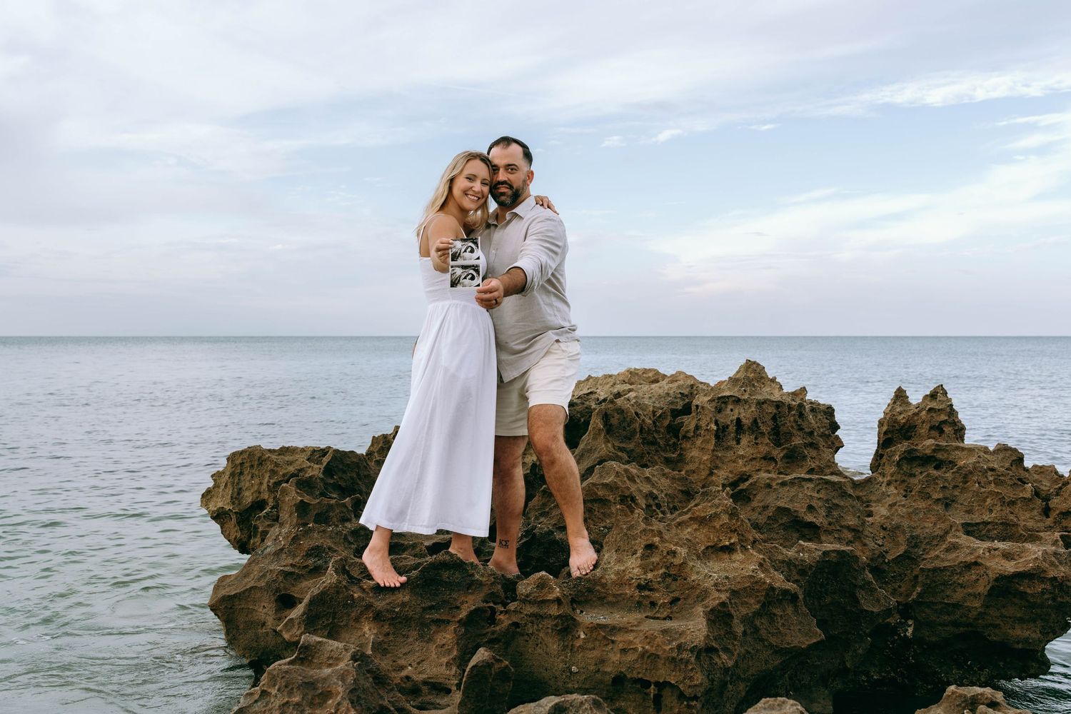 Couple embracing on rocky shoreline with ocean backdrop, woman in white dress, man in casual attire.