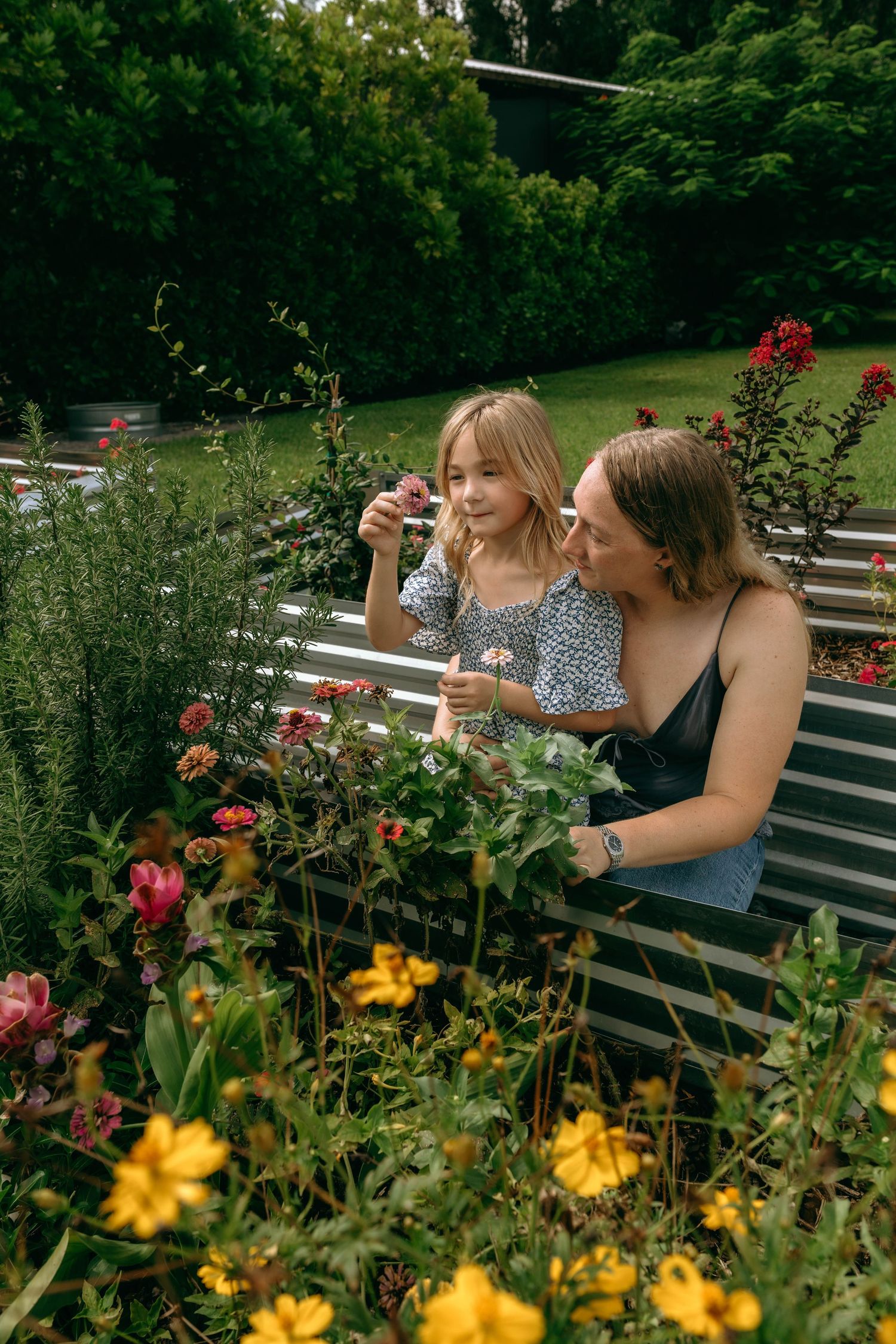 A woman and young girl admire colorful flowers in a lush garden surrounded by blooming plants and greenery.