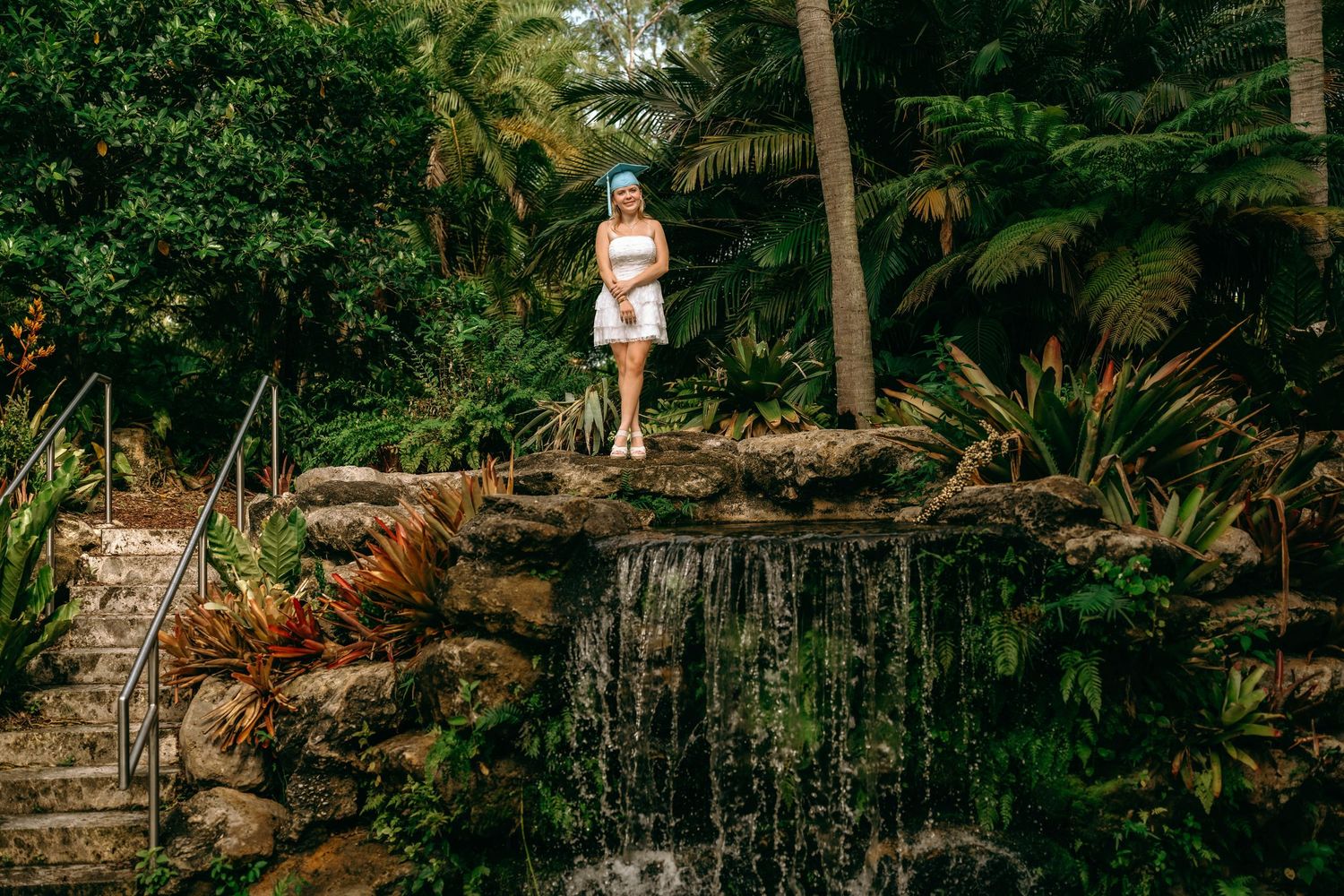 Woman in white dress standing on rocky waterfall surrounded by lush tropical greenery and palm trees.