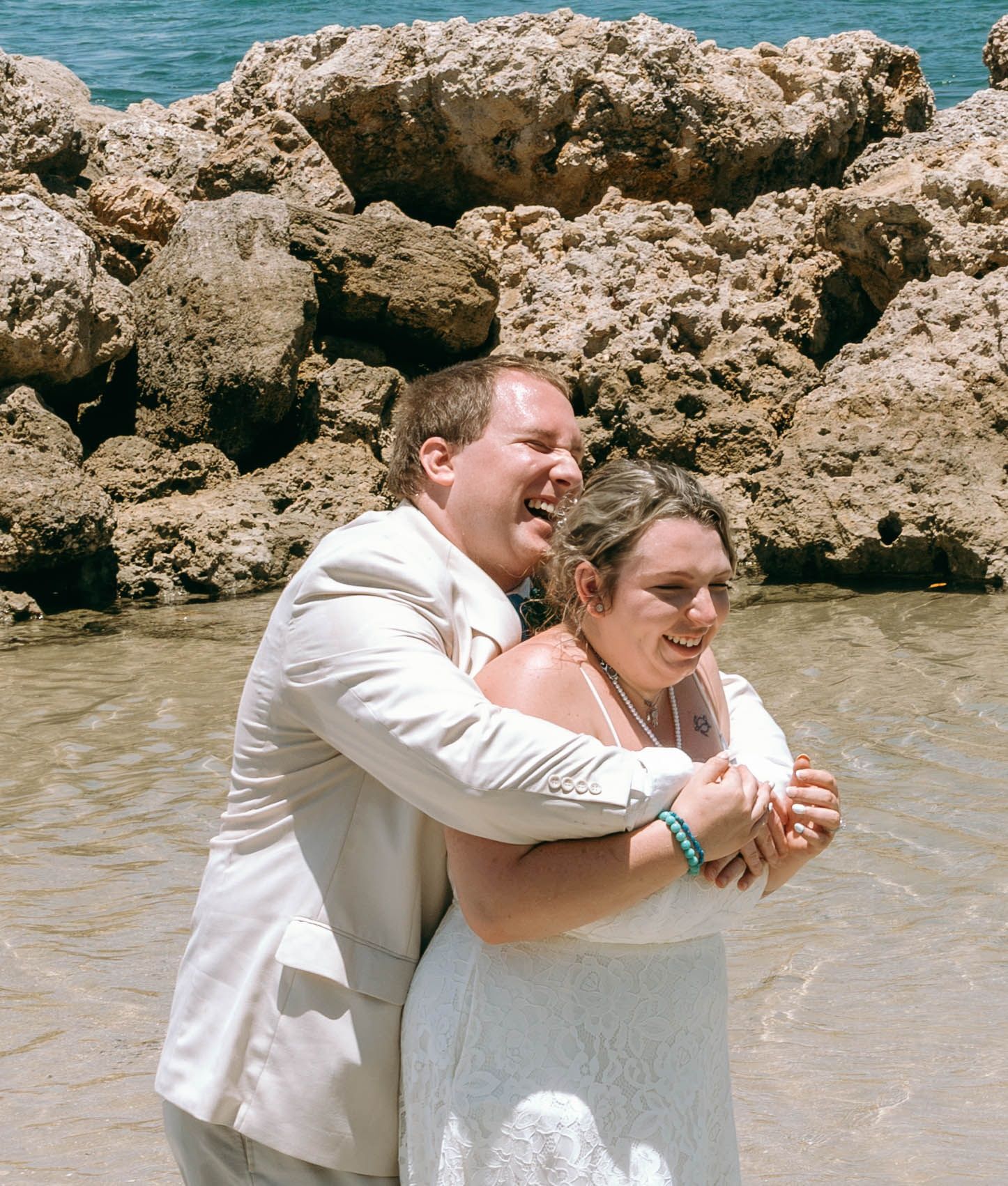 Couple in wedding attire embracing and laughing on a rocky beach shoreline, groom in white suit, bride in white dress.